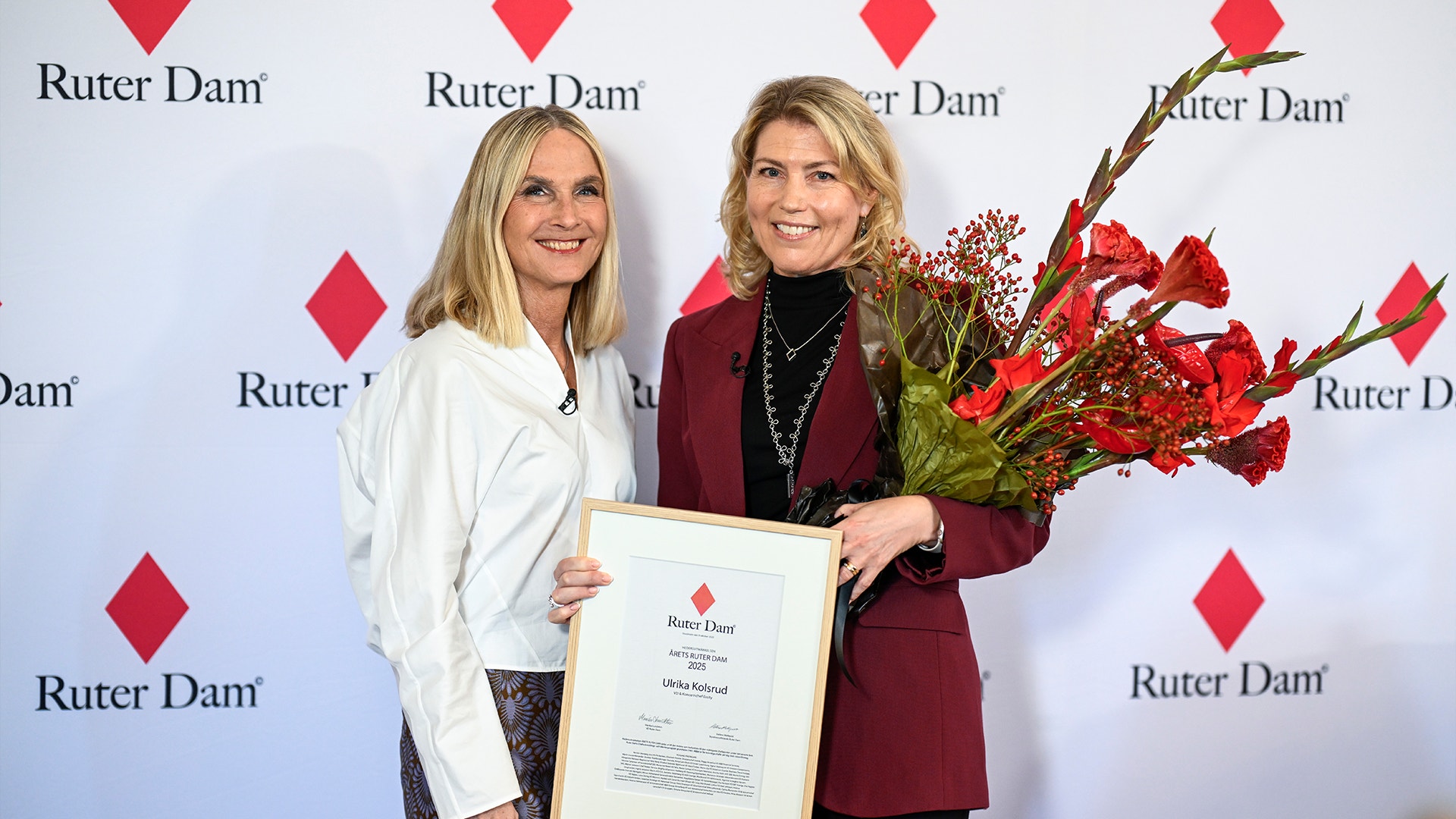 Två kvinnor vid ett ceremoniellt event, en i vit blazer och en i vinröd blazer, håller ett diplom och en bukett röda blommor framför en Ruter Dam-bakgrund.