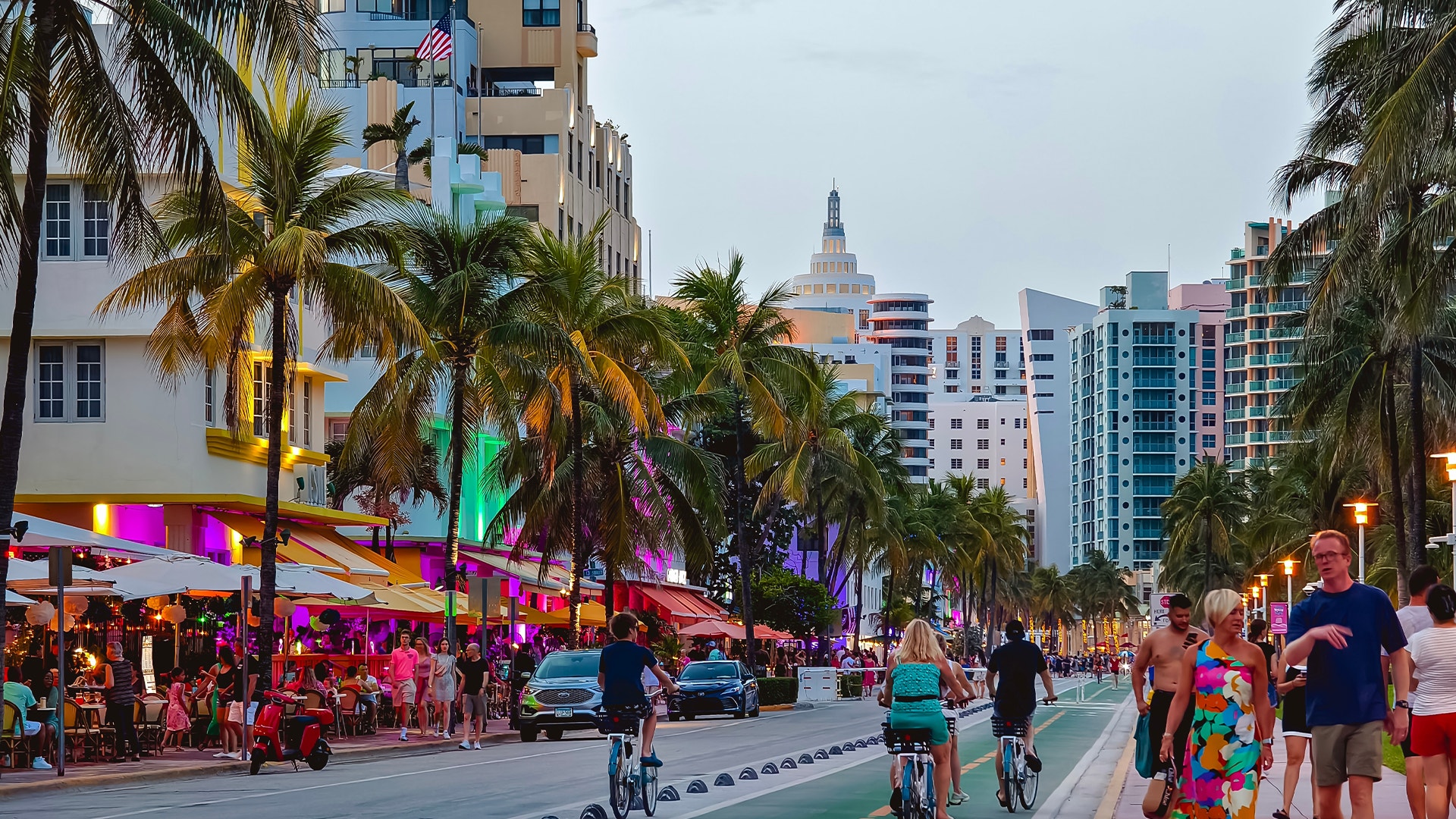 Miami Beach Ocean Drive på kvällen med palmträd, färgglada byggnader, neonljus och människor som promenerar och cyklar längs gatan.