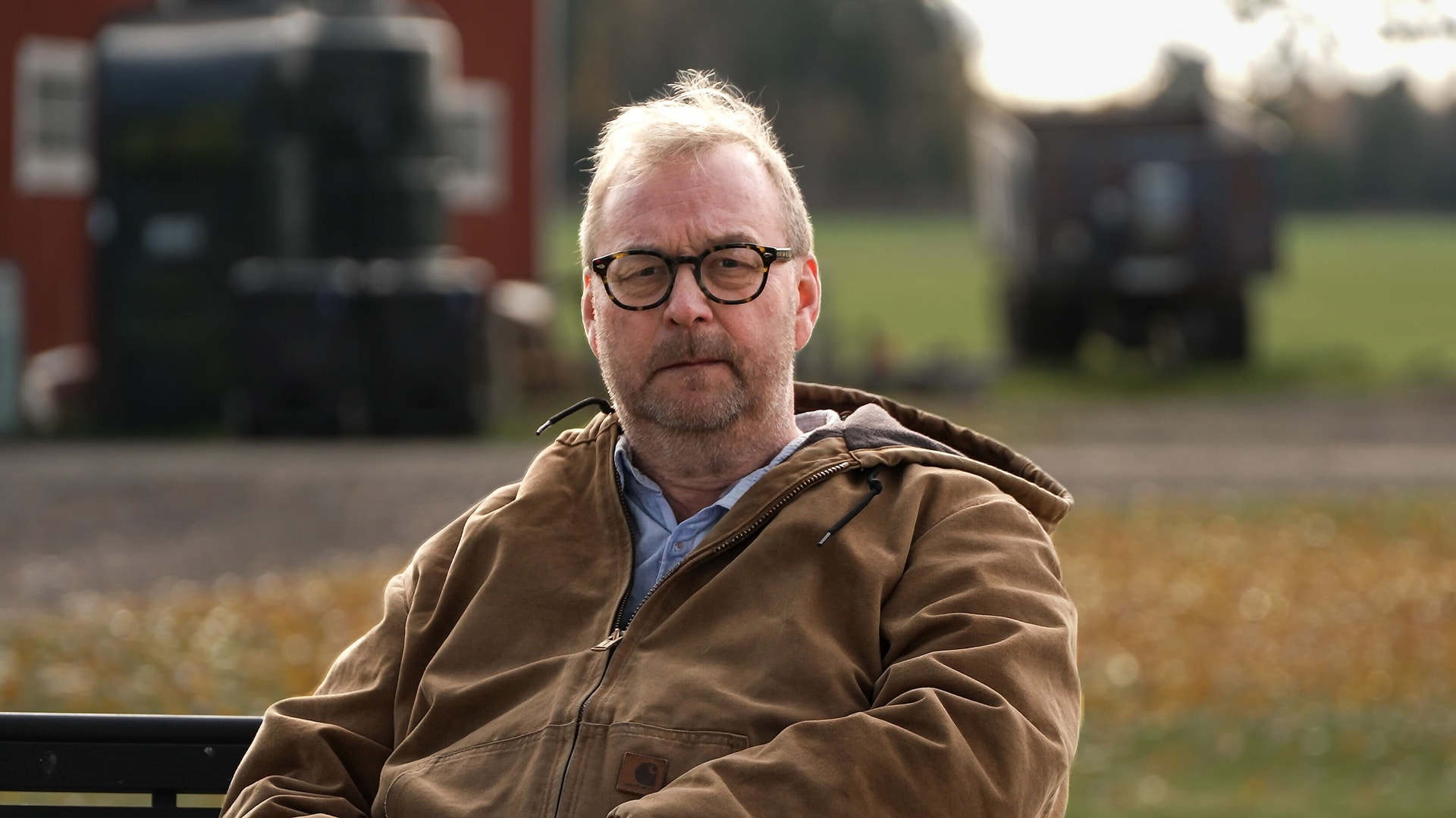 Porträtt av en man med glasögon i brun jacka framför jordbruksmaskiner