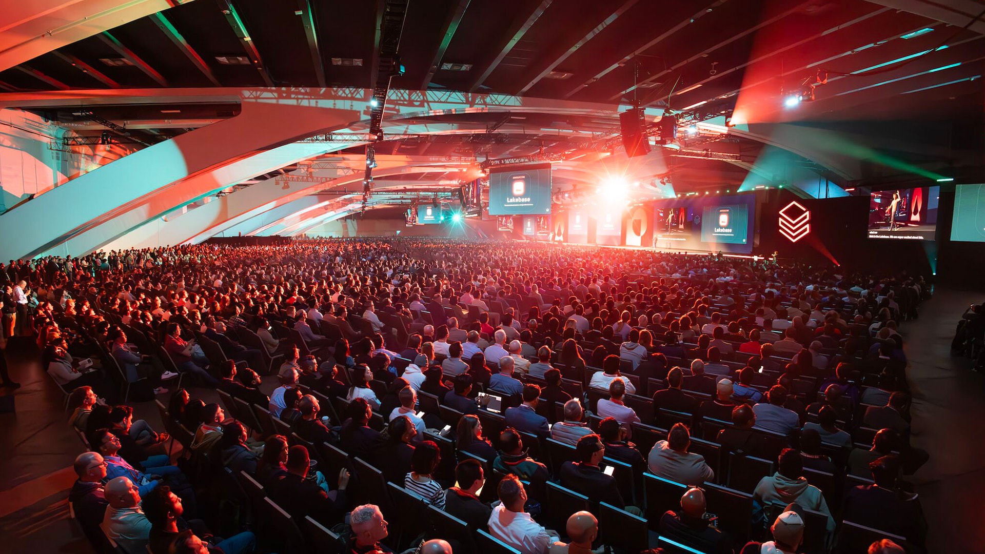 En stor konferenssalong fylld med hundratals åhörare under röd och turkos belysning, med en scen synlig i bakgrunden.