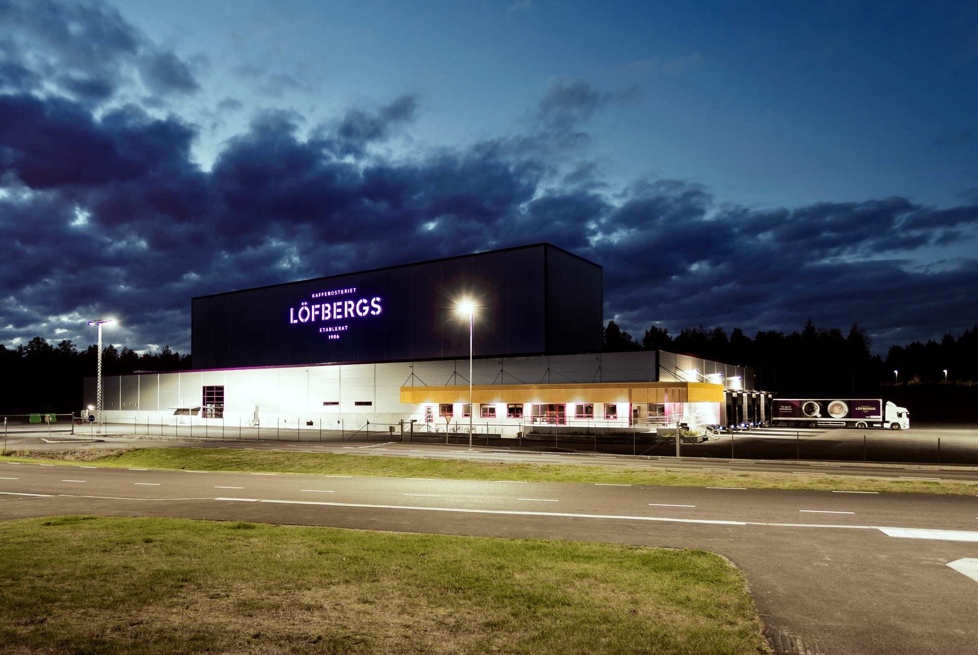 Löfbergs-byggnaden fotograferad vid skymning. En modern industribyggnad med svart tak och vita väggar, belyst av strålkastare. Vägen och gräsmattan framför byggnaden syns under en dramatisk himmel med moln.