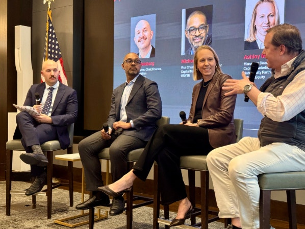Fyra personer sitter på scenen under en paneldiskussion. I bakgrunden visas porträtt av andra personer på en skärm, och en amerikansk flagga är synlig till vänster.