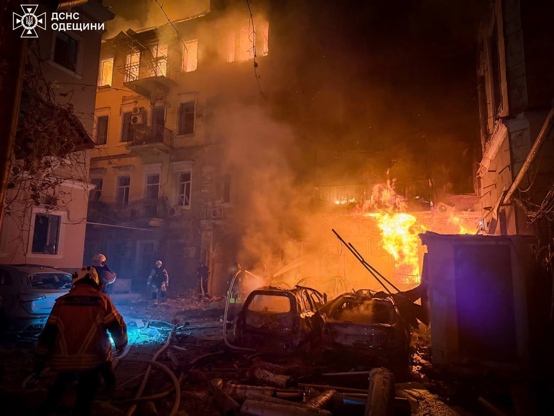 En stor brand rasar på en stadsgata på natten. Intensiva orange och gula flammor sprider sig från fordon och byggnader. Brandmän i röda uniformer arbetar för att bekämpa branden. Tjock rök täcker området runt en flervåningsbyggnad.