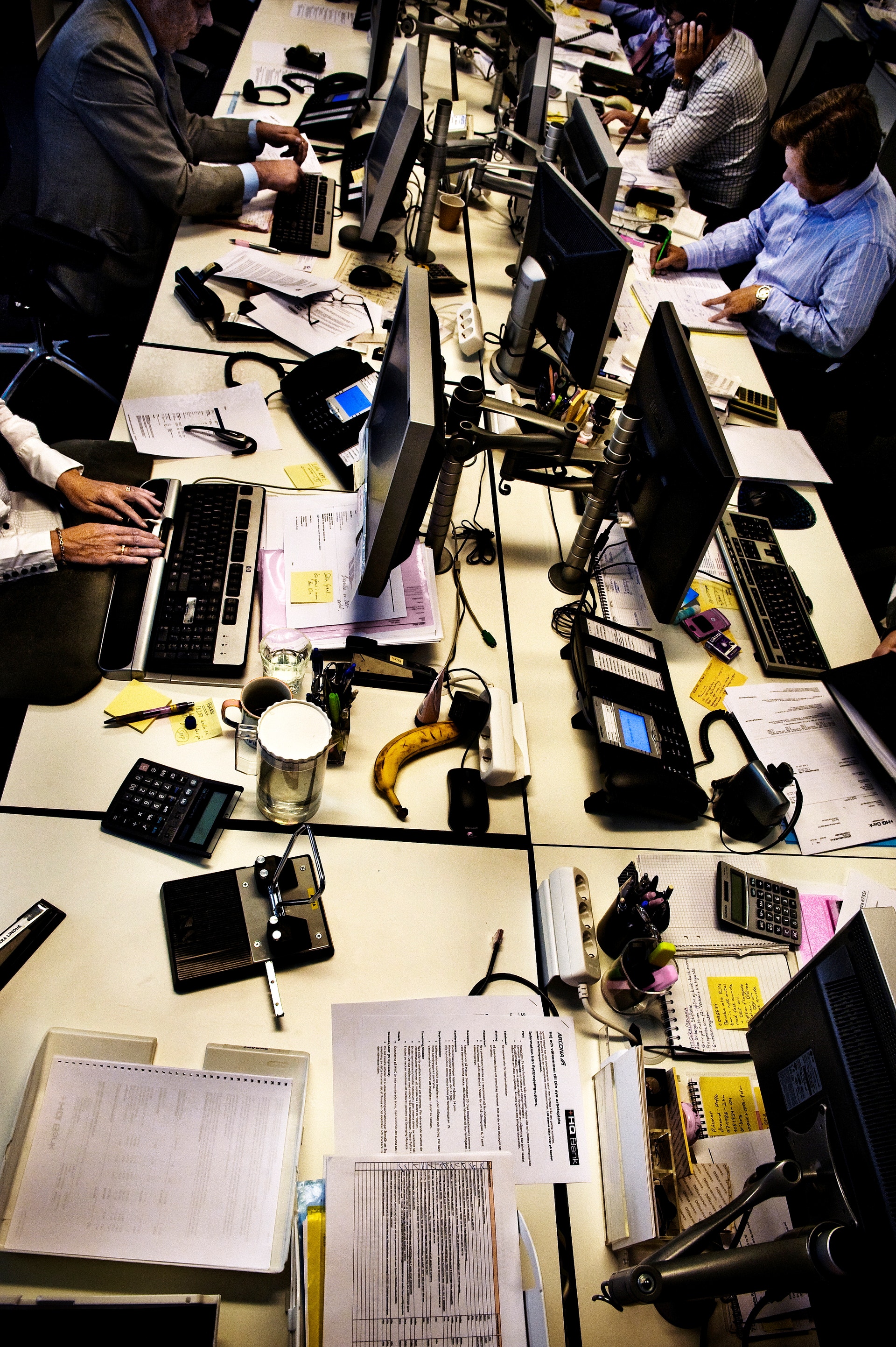 Flera personer arbetar tillsammans vid datorer och skrivbord i ett öppet kontorslandskap. Överblicksbild från ovan visar laptops, dokument och arbetsverktyg på borden.