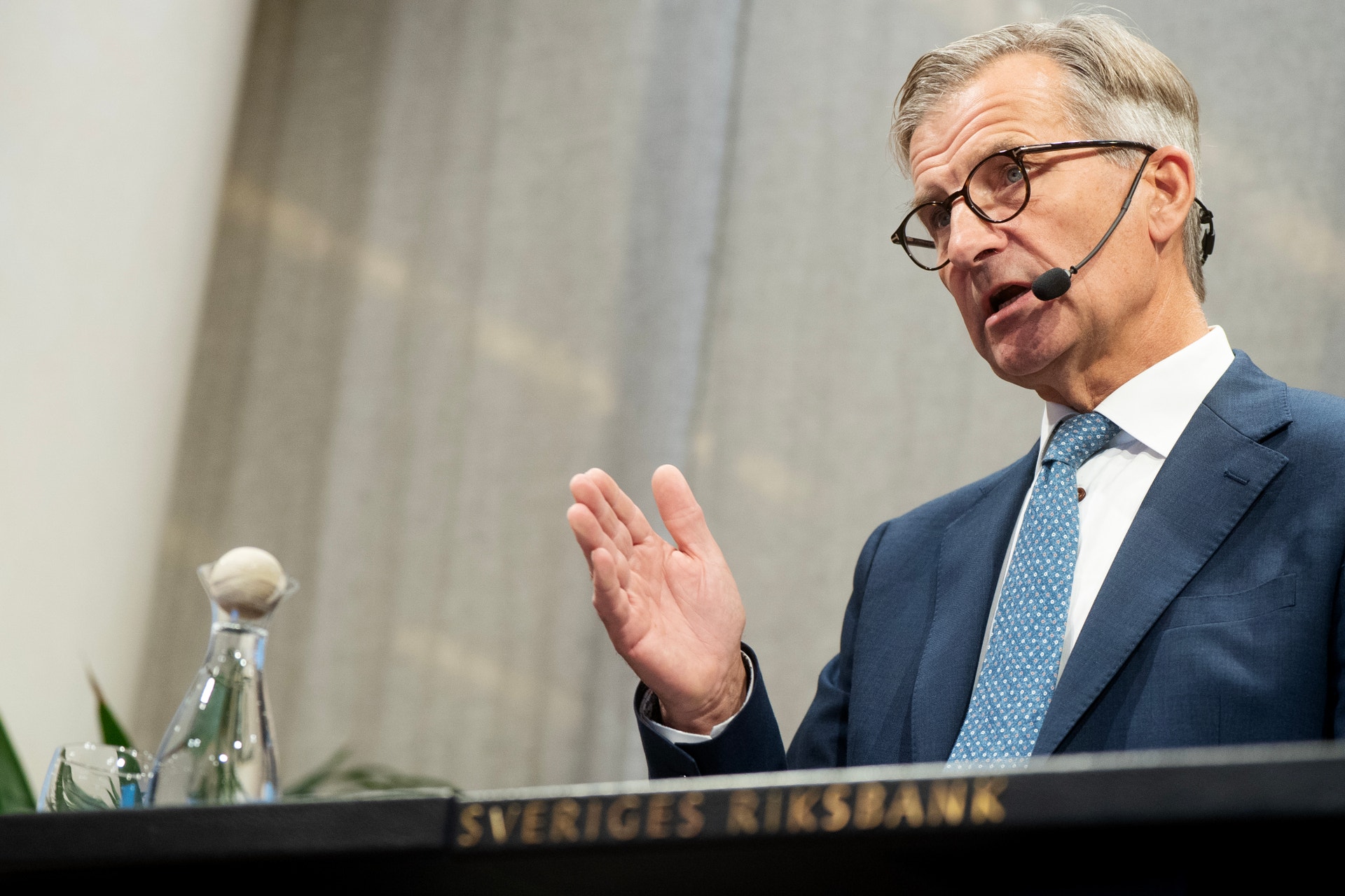 Man i blå kostym och glasögon talar vid podium märkt Sveriges Riksbank, med mikrofon och gestikulerande hand.