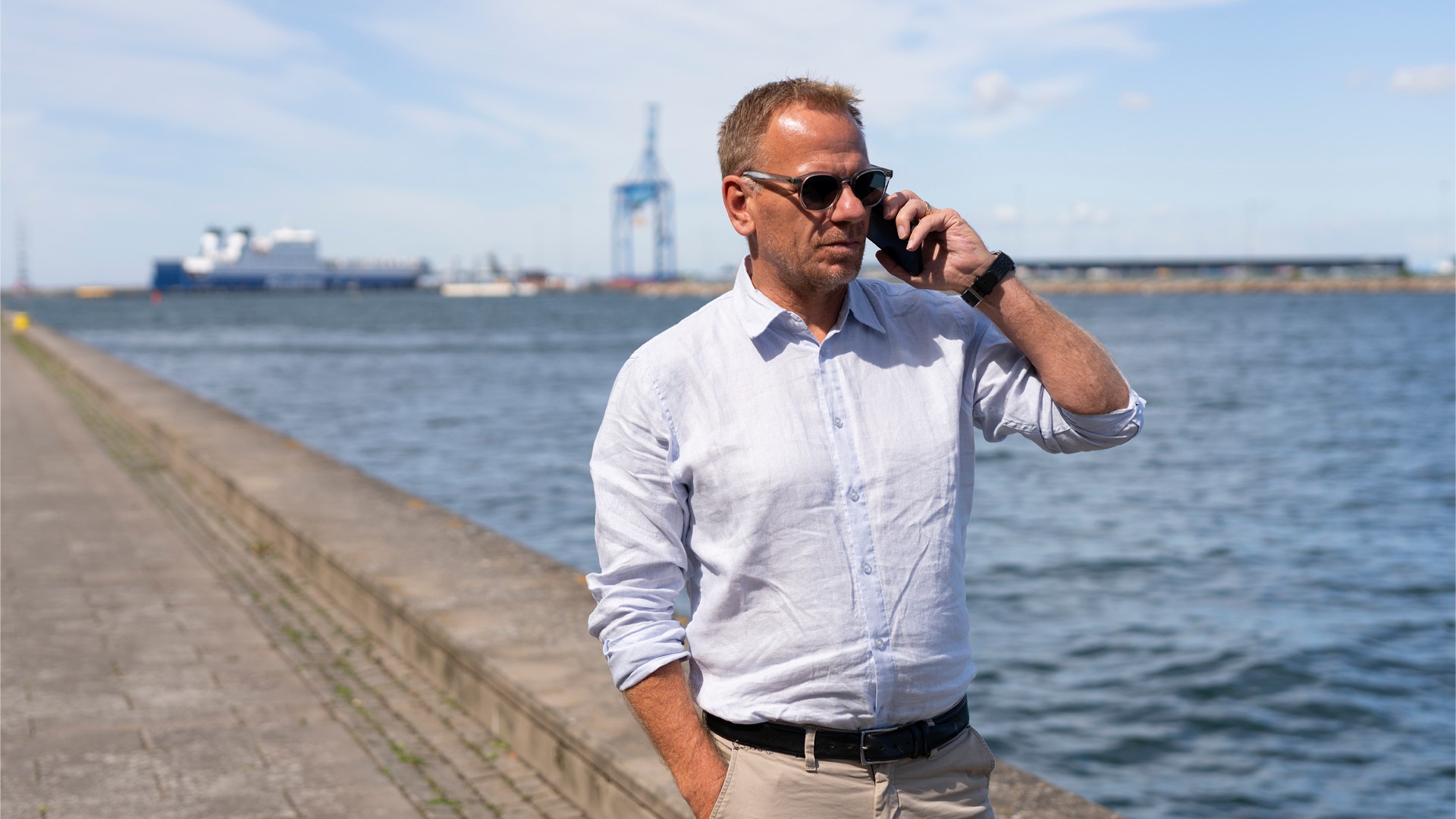 Man i vit skjorta och solglasögon talar i telefon på en kaj vid hamnen med fartyg i bakgrunden.