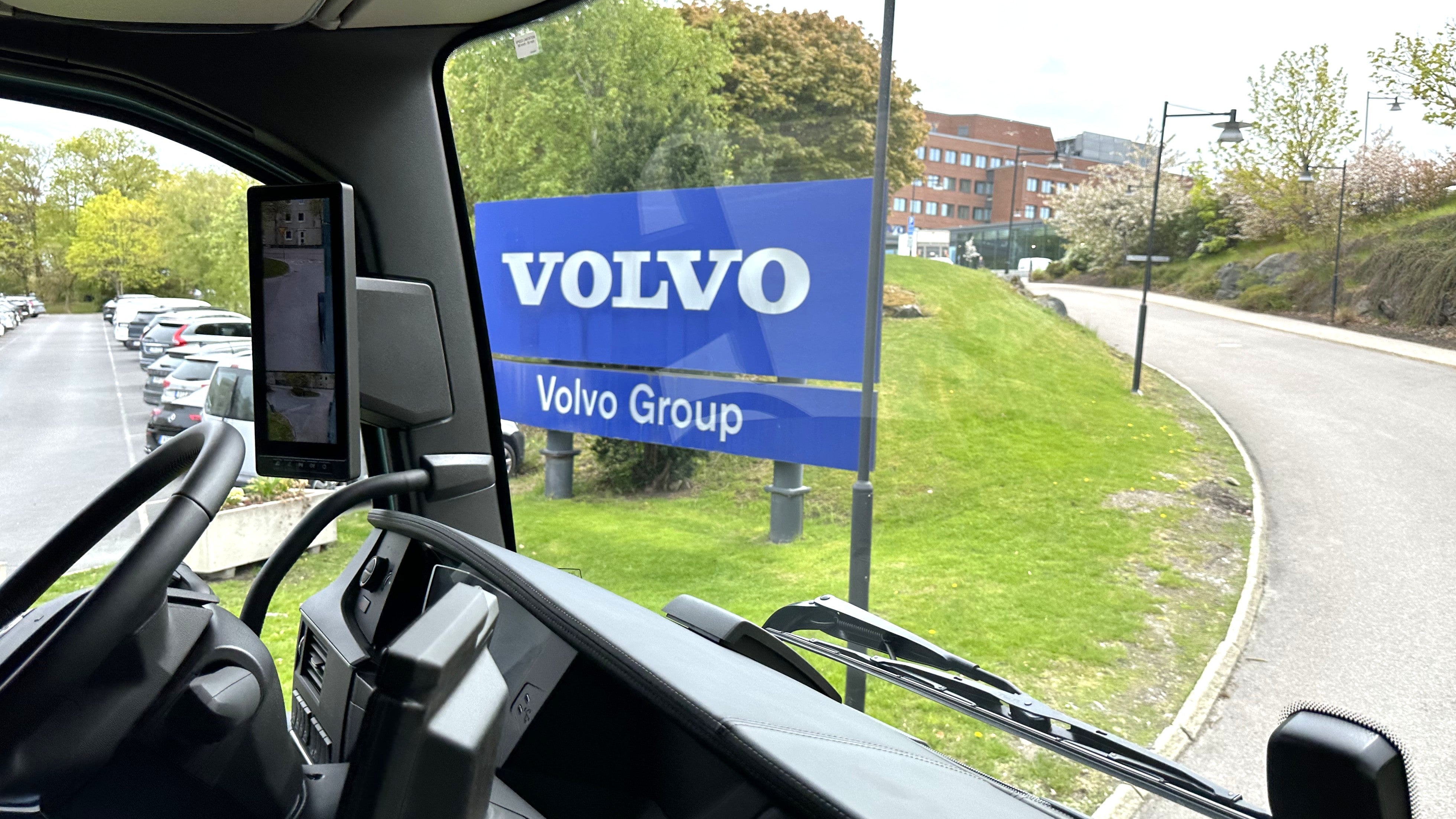 Utsikt från ett fordon mot en blå Volvo Group-skylt med industriell miljö och gröna områden i bakgrunden.