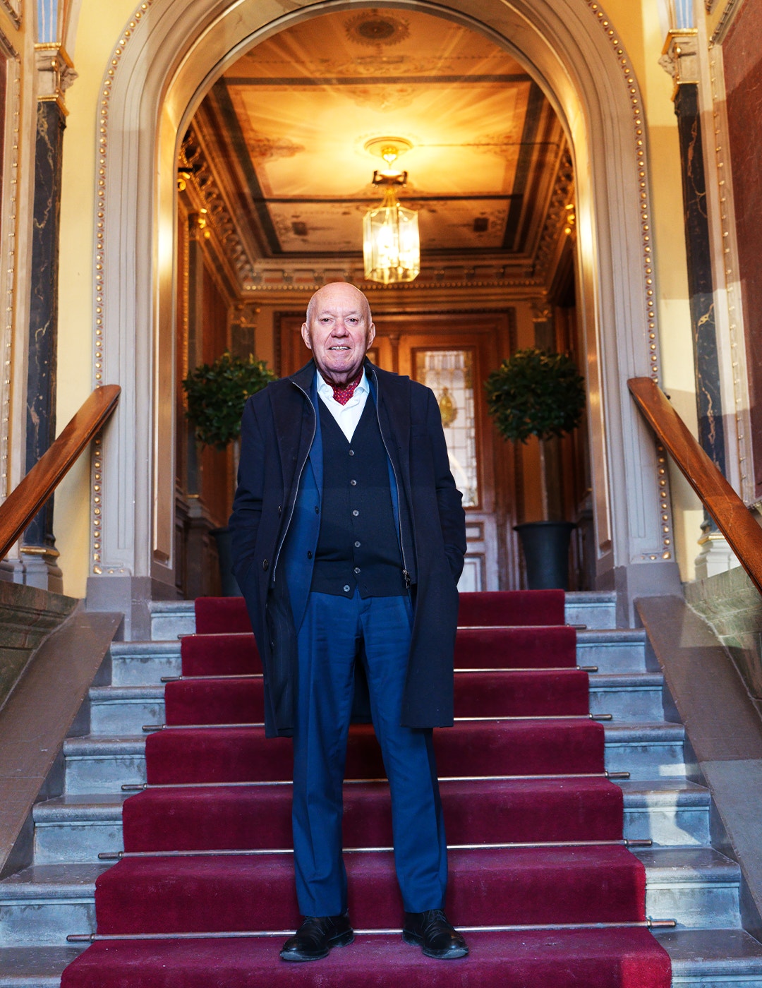 En man står på en röd matta på en stor marmortrappa i en elegant klassisk interiör med gyllene dekorationer och en glaskrona.