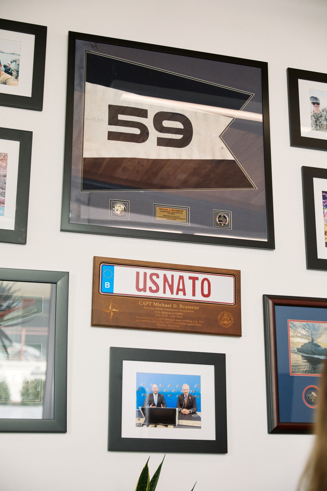 Vägg med inramade minnessaker och plaketter, inklusive ett nummer 59-skyltar och en USNATO-plakette bland andra ramar.