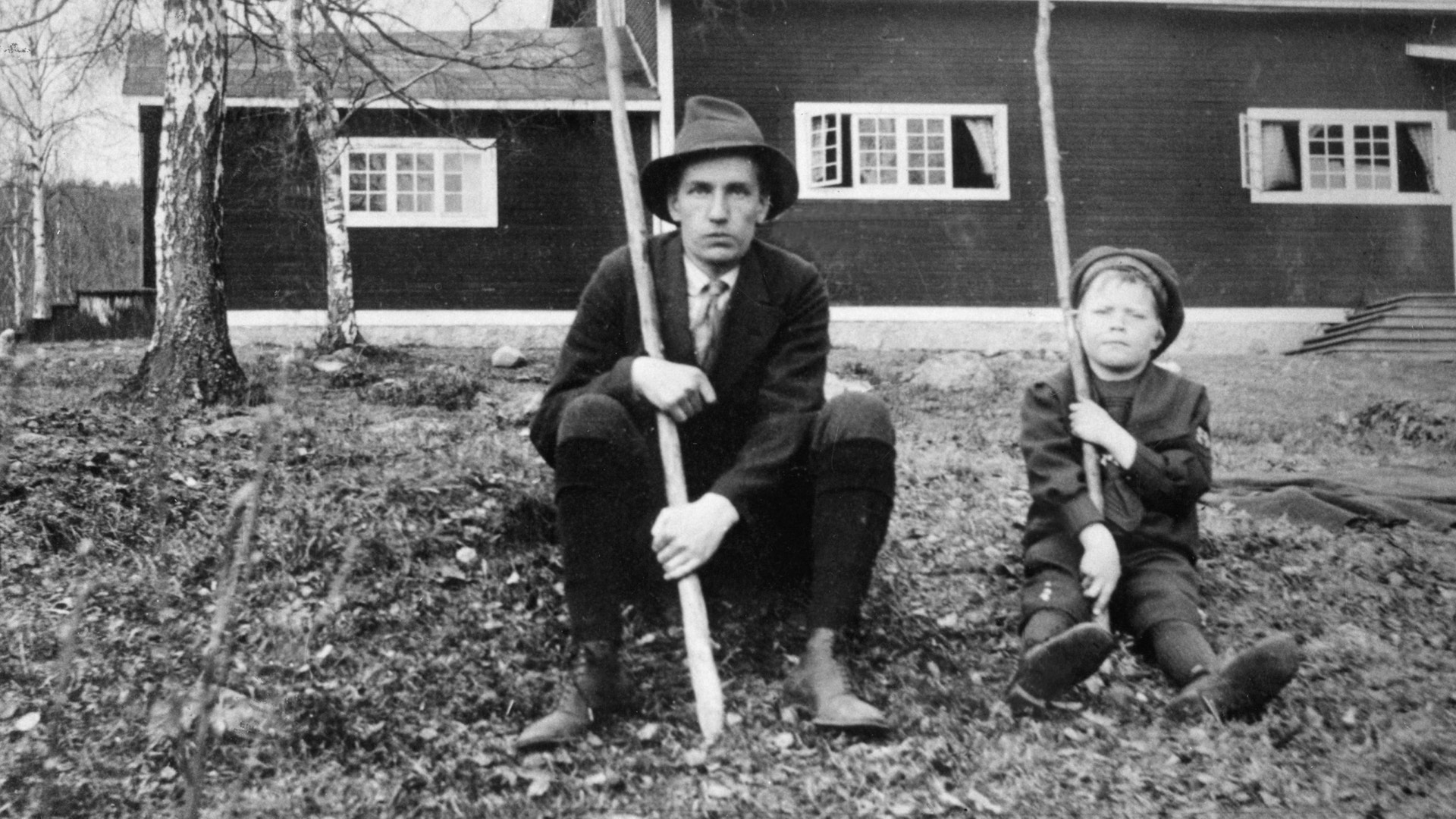 Svartvitt äldre foto av en man och en pojke som sitter på marken framför ett hus, båda håller i långa käppar.