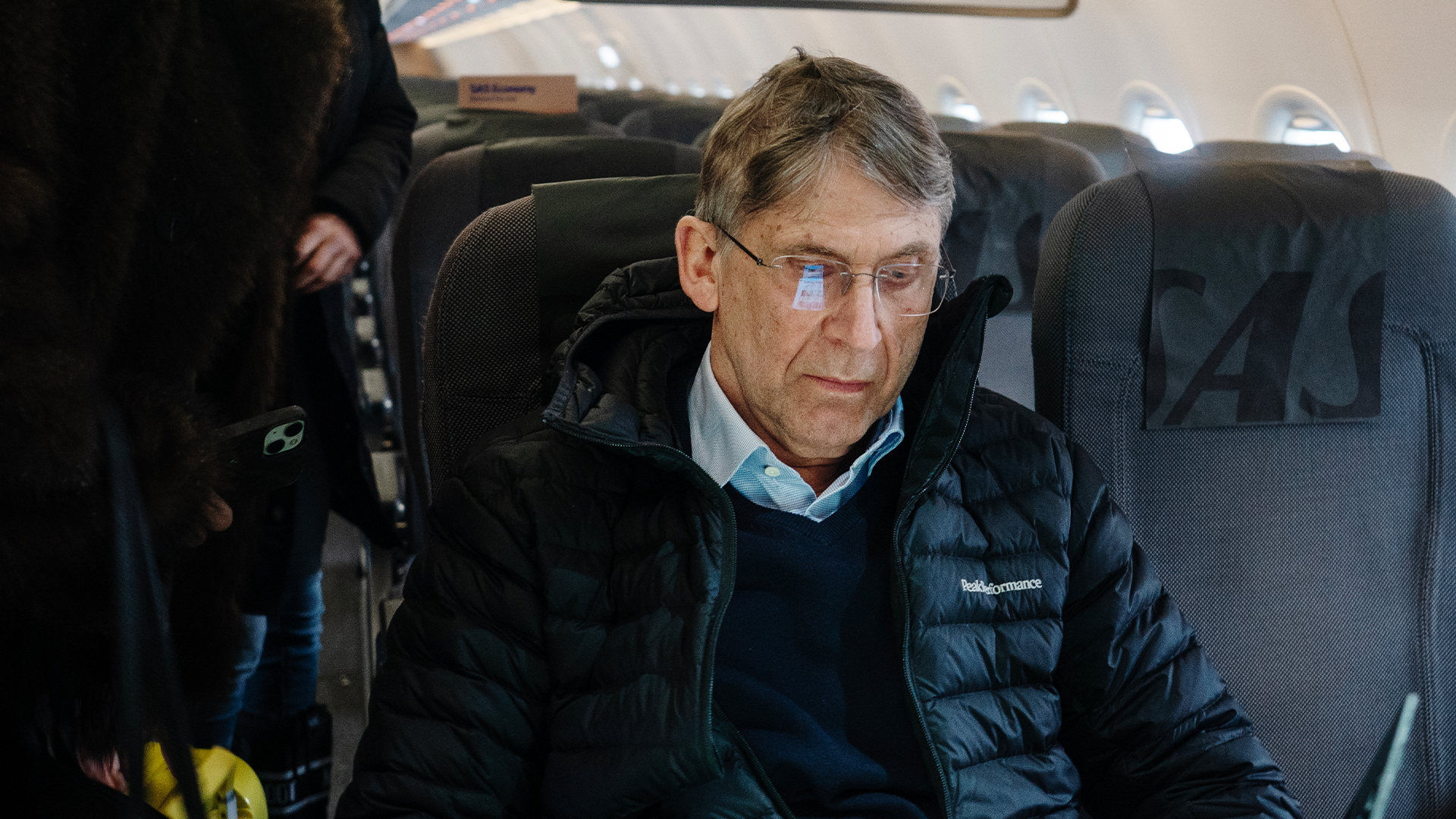 Man sitter i ett flygplan och bär svart dunjacka och ljusblå ansiktsmask