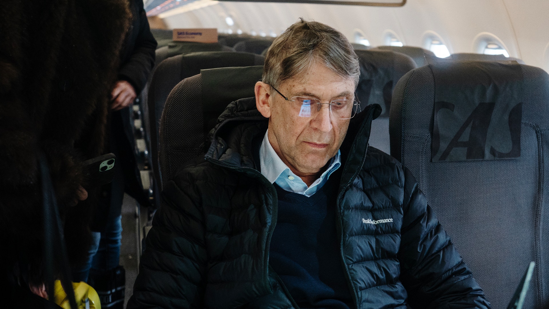 Man sitter i ett flygplan och bär svart dunjacka och ljusblå ansiktsmask