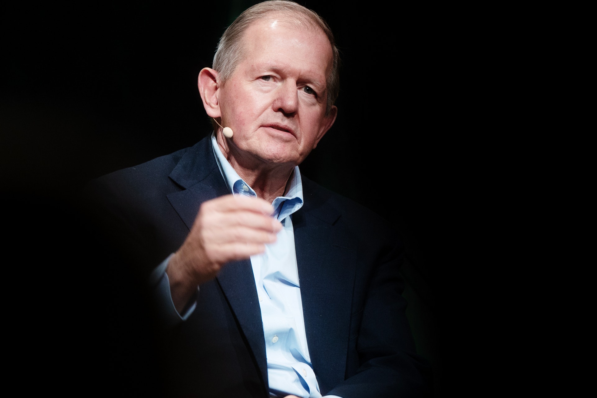 En äldre man i mörkblå kostym och ljusblå skjorta gestikulerar medan han talar mot en mörk bakgrund.