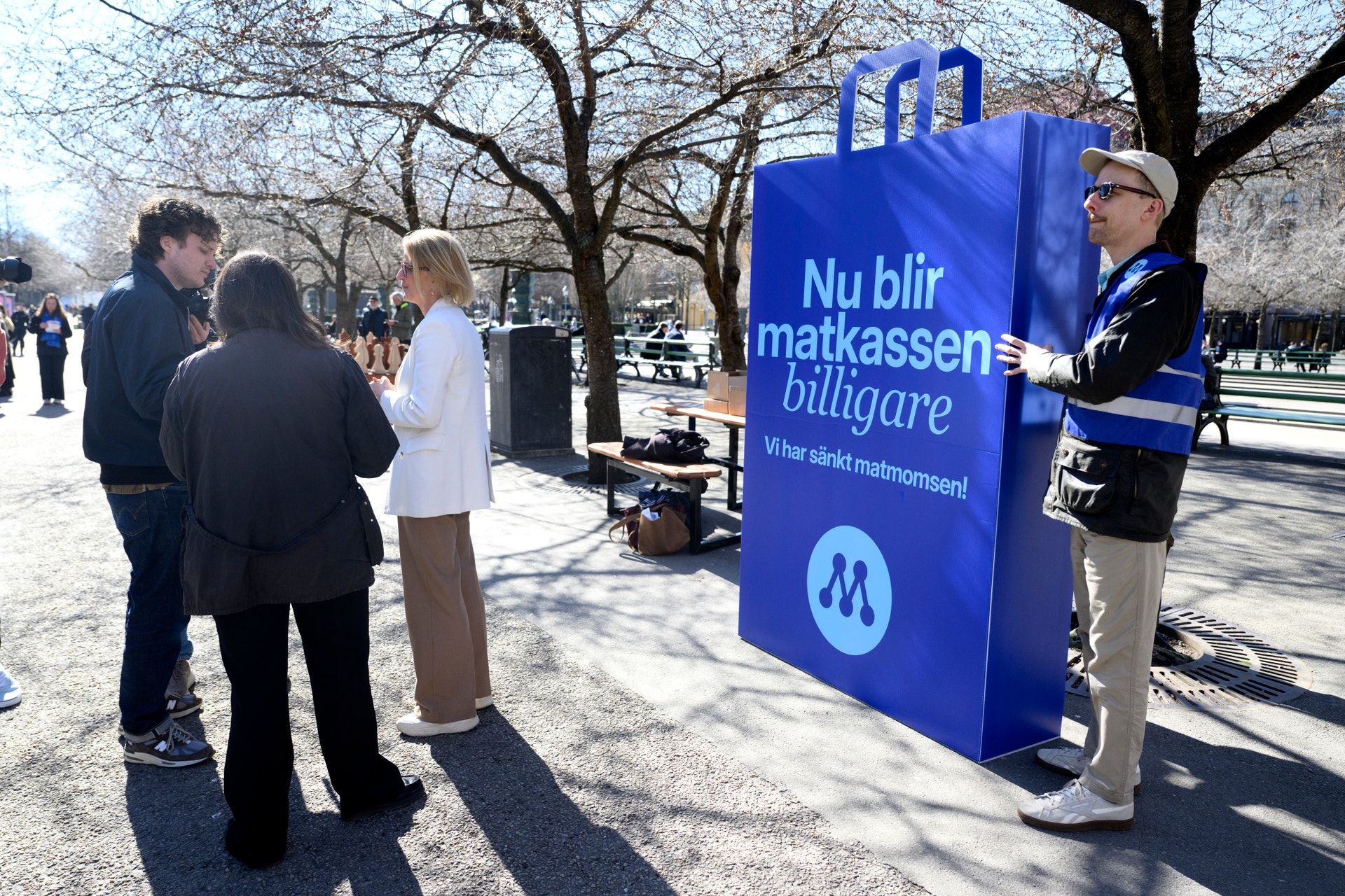 En stor blå skylt vid en utomhusevent där människor står samlade. Skyltens text säger 'Nu blir matkassens biligare' med ett M-logga.