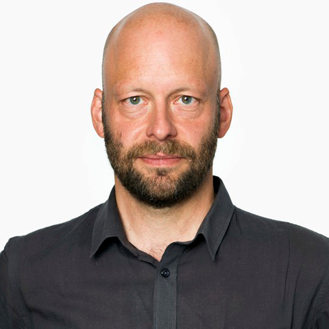 Porträtt av en man med skägg och svart skjorta mot vit bakgrund.