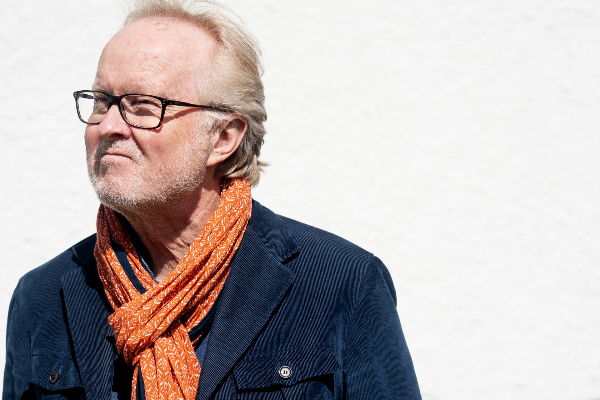 Porträtt av en äldre man med glasögon och vit hår, klädd i mörkblå jacka och orange scarf.