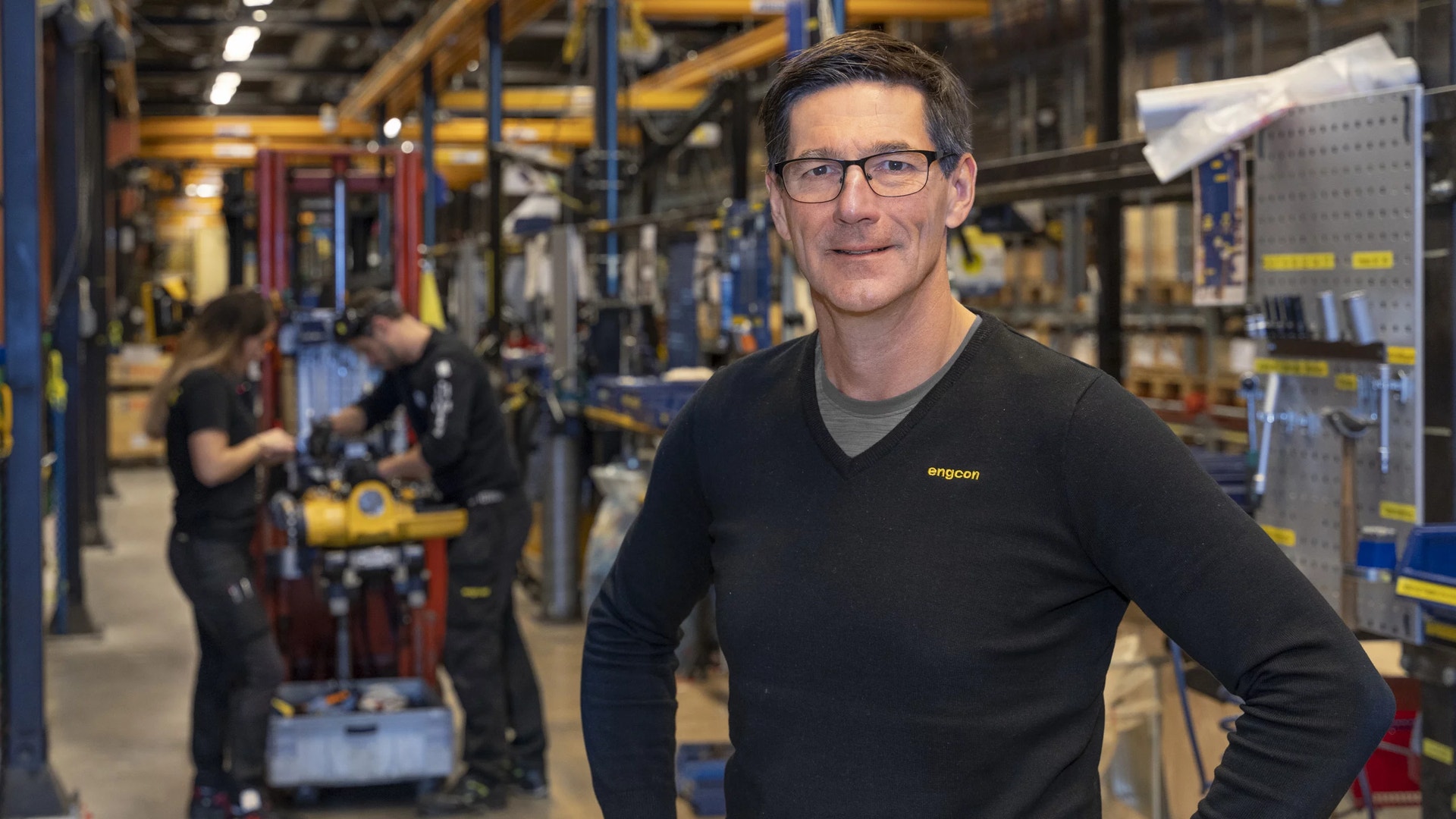 Man i svart skjorta står i en industriell verkstad med maskiner och utrustning i bakgrunden.