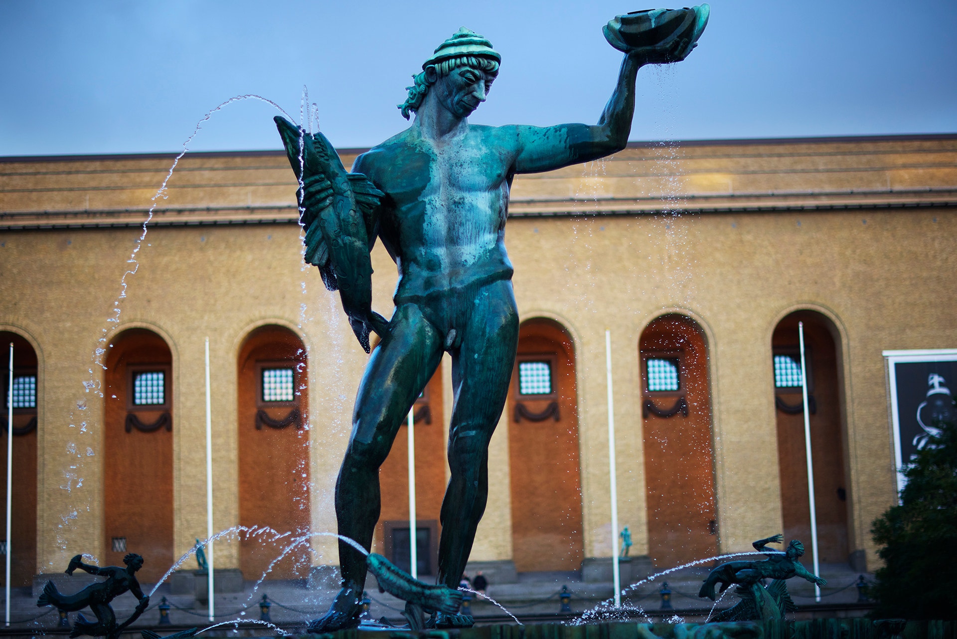 Grön bronsstaty av en man som håller upp en snäcka, framför en gul byggnad med välvda fönster. Vatten sprutar runt statyn.