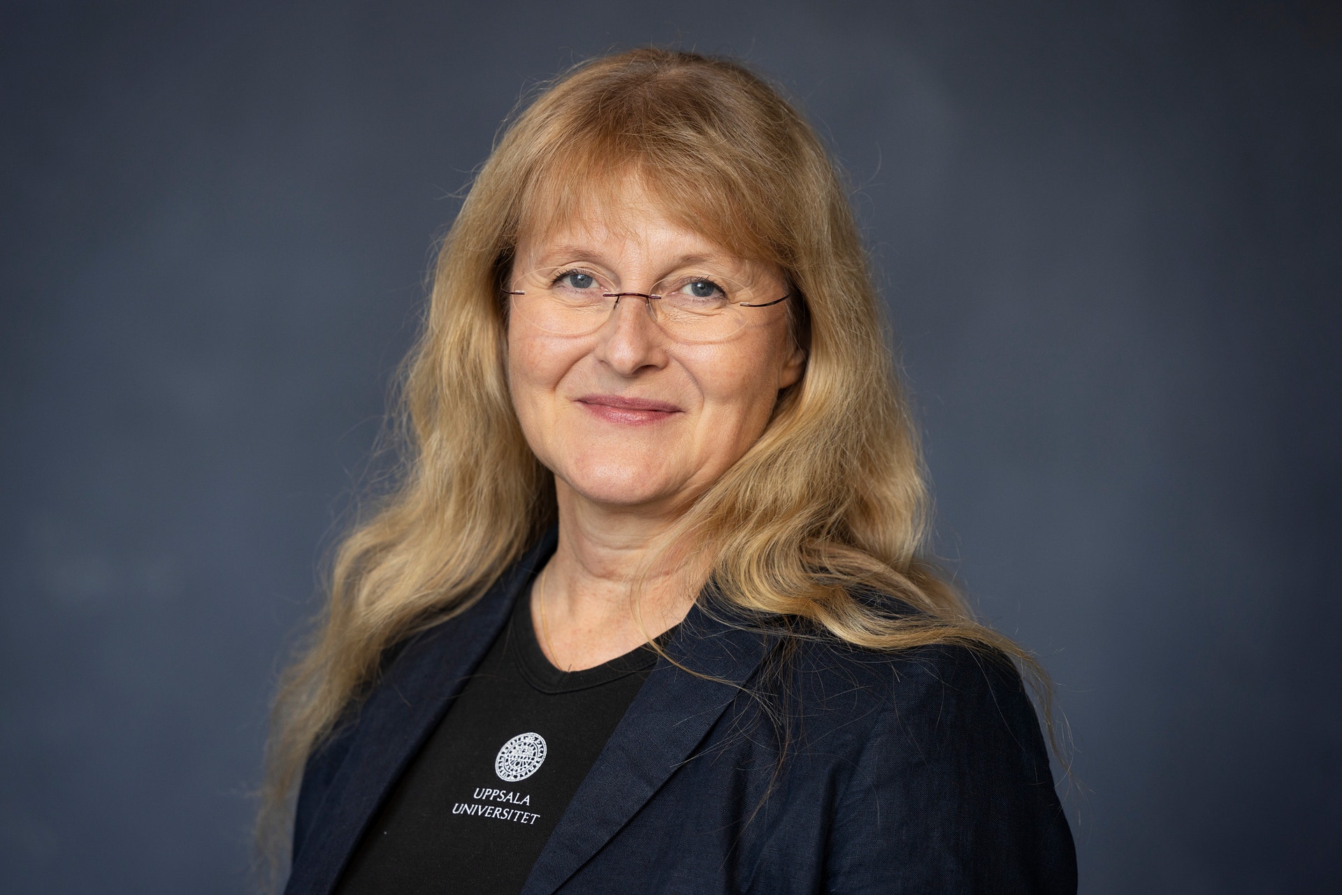 Porträtt av en leende kvinna med långt ljust hår och glasögon, klädd i mörk kavaj och svart tröja med Uppsala universitets logotyp.