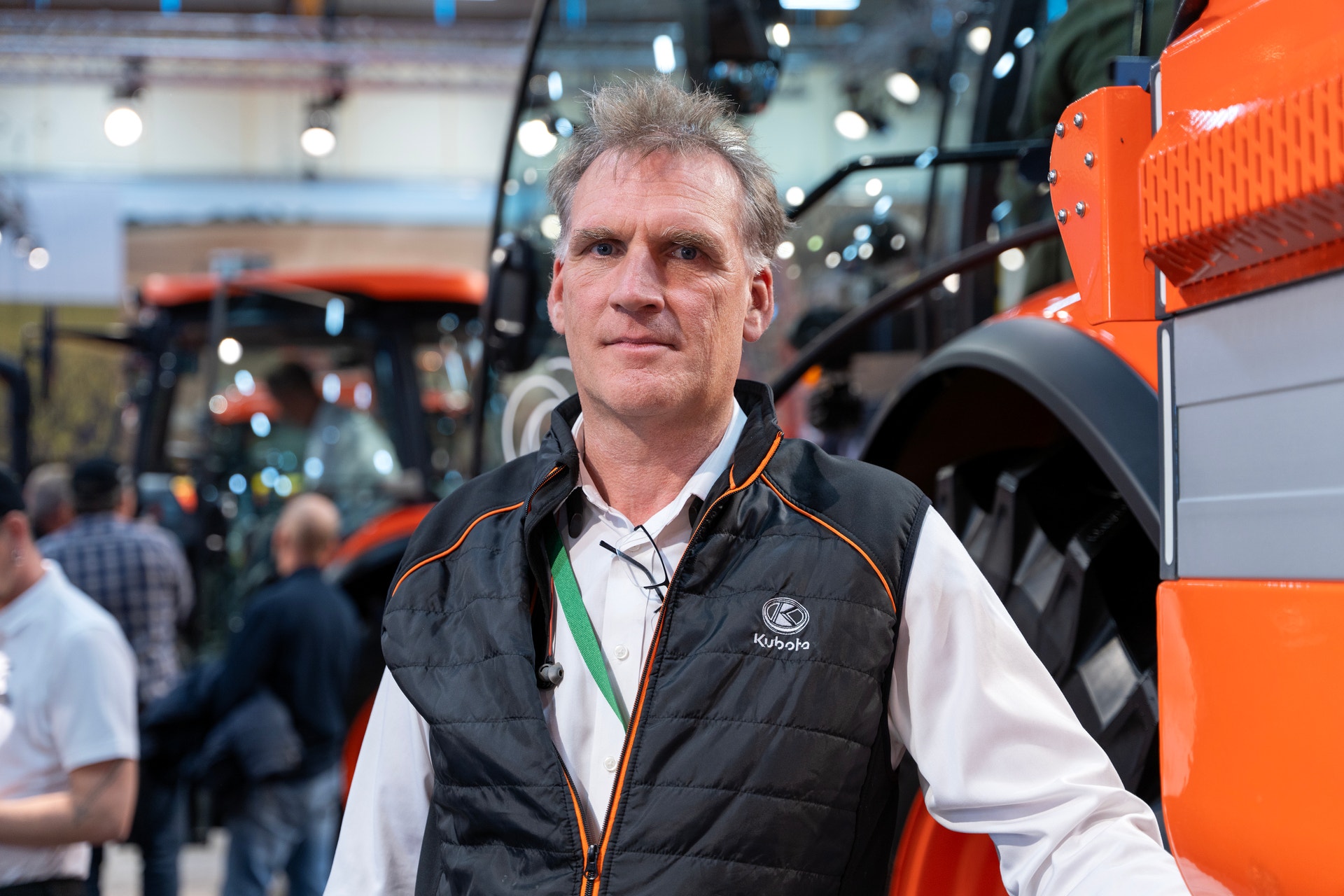 Man i svart väst och vit skjorta tittar in i kameran framför en orange traktor på en lantbruksmässa.