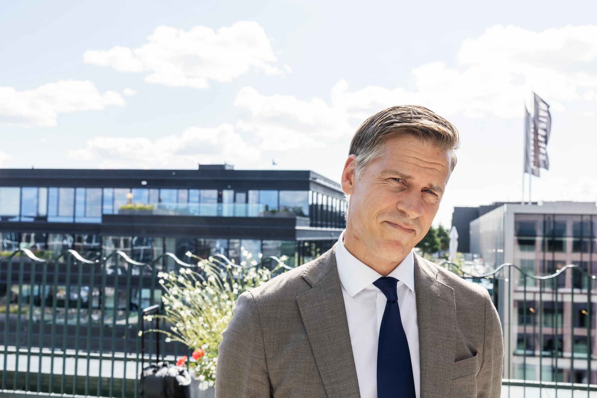 Porträttfoto av en man i grå kostym och mörkblå slips som står utomhus framför moderna byggnader under blå himmel med vita moln.