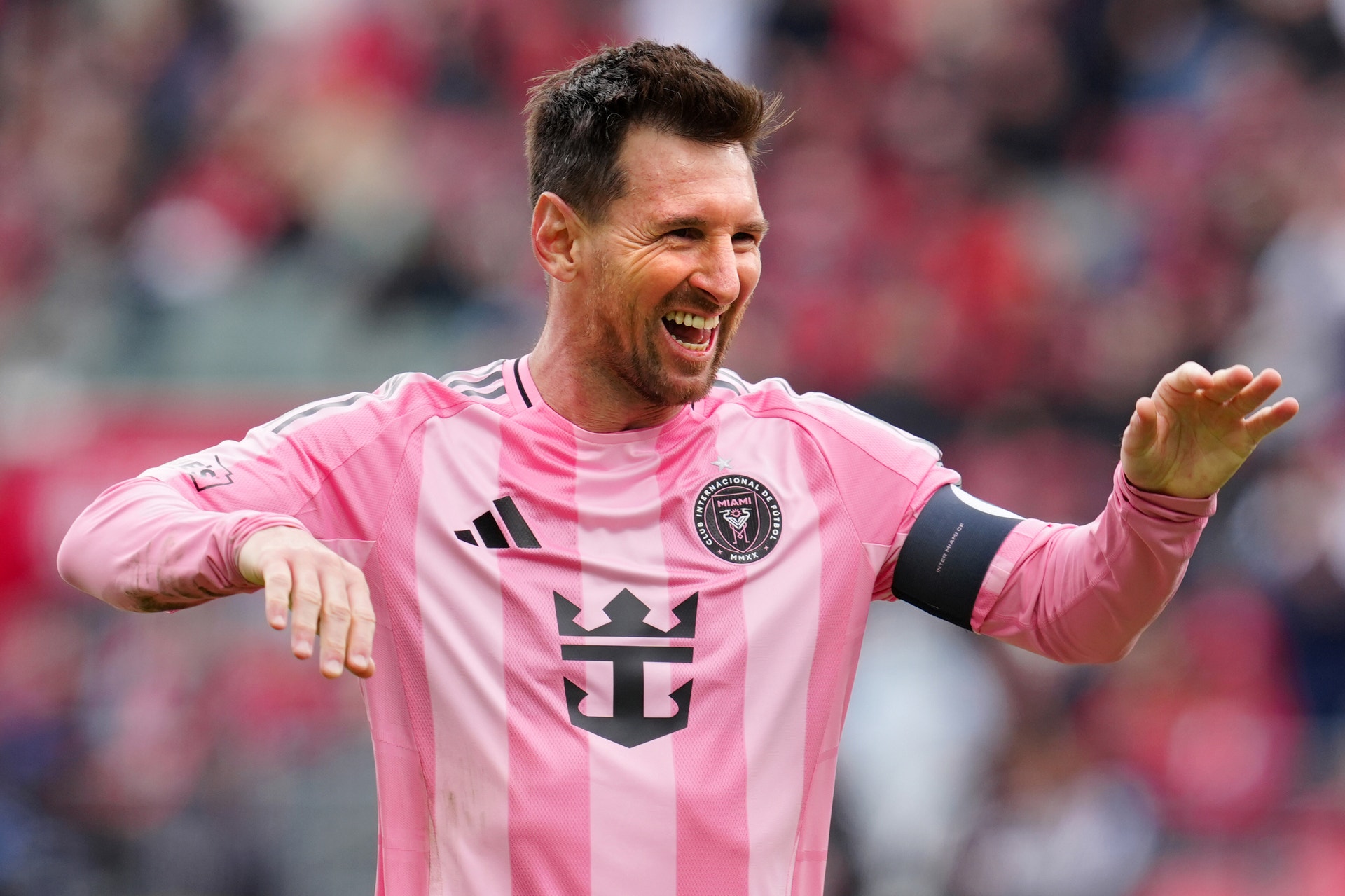 Fotbollsspelare i rosa lag-tröja ler och gestikulerar under en match med publik i bakgrunden.