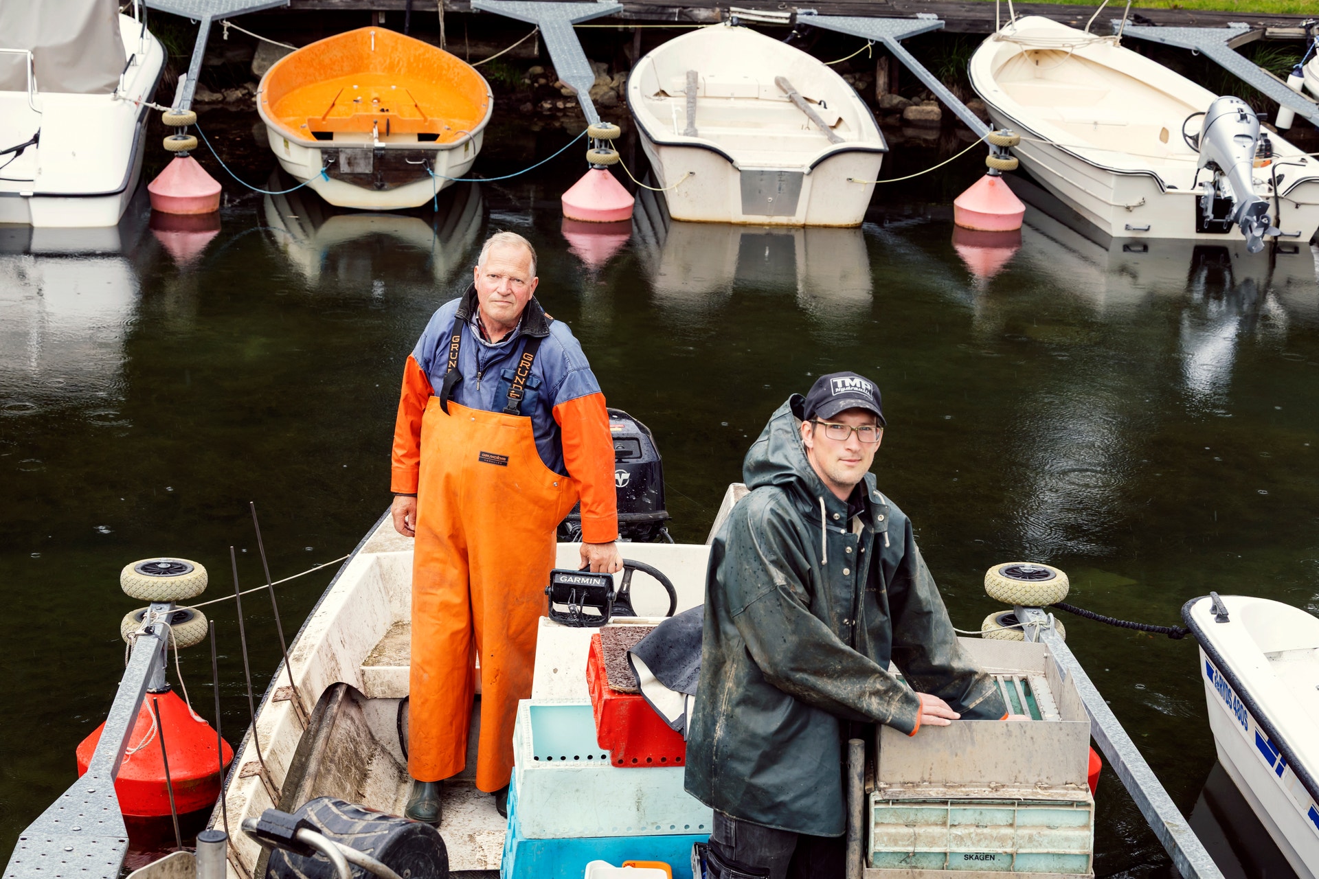 Två personer står i en fiskebåt vid en brygga. Personen till vänster bär en orange fiskardräkt och personen till höger bär en svart jacka och keps. I bakgrunden syns flera andra båtar förtöjda vid bryggan.