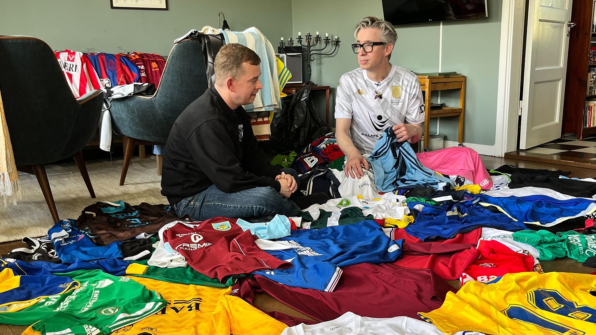 Två personer sitter på golvet och sorterar en stor samling färgglada sportkläder och tröjor i olika färger.