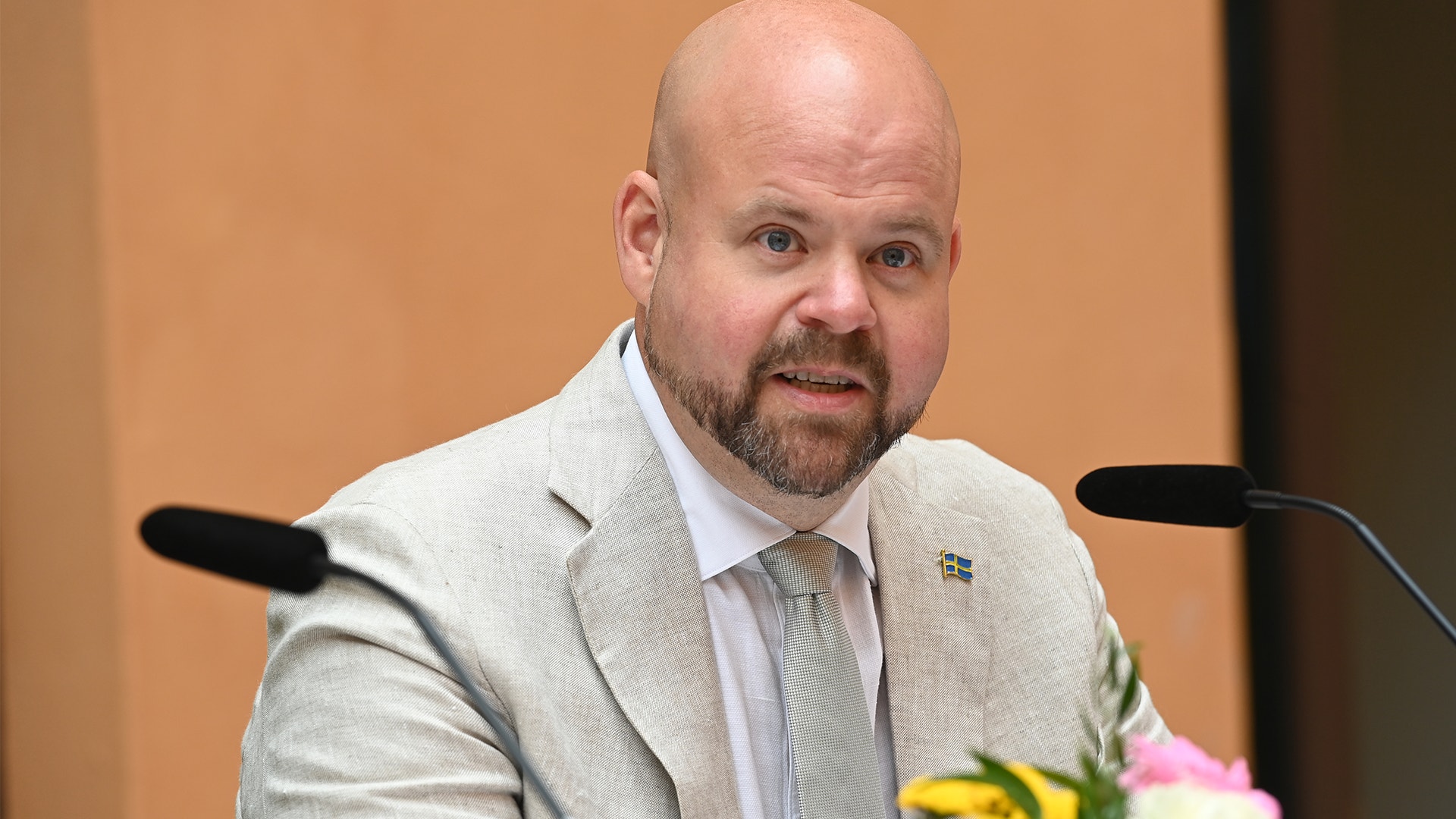 Porträtt av en man i ljus blazer och slips som sitter vid ett bord framför mikrofoner under ett möte eller en presentation.