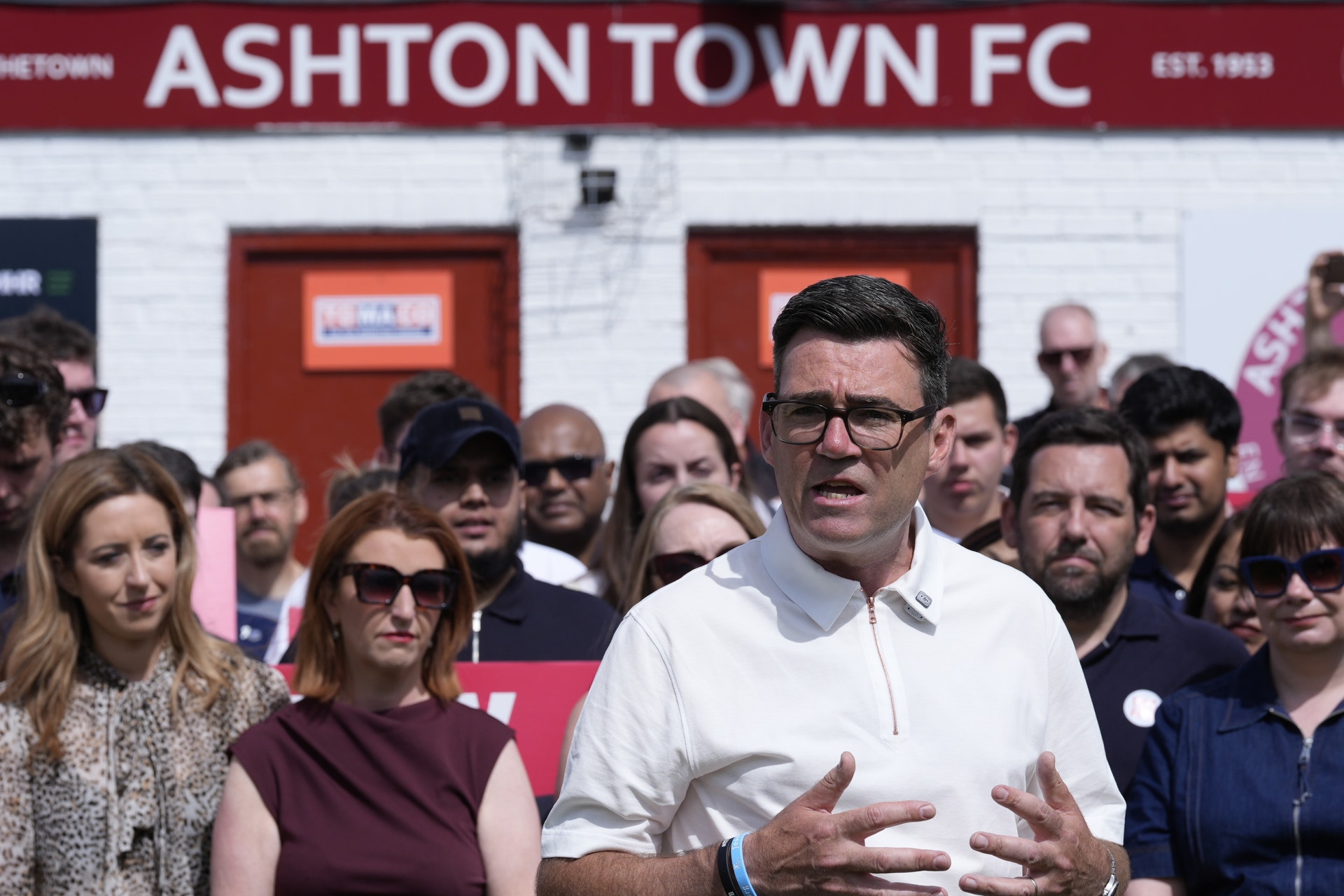 En man i vit skjorta talar inför en grupp människor utanför Ashtontown FC-stadion.