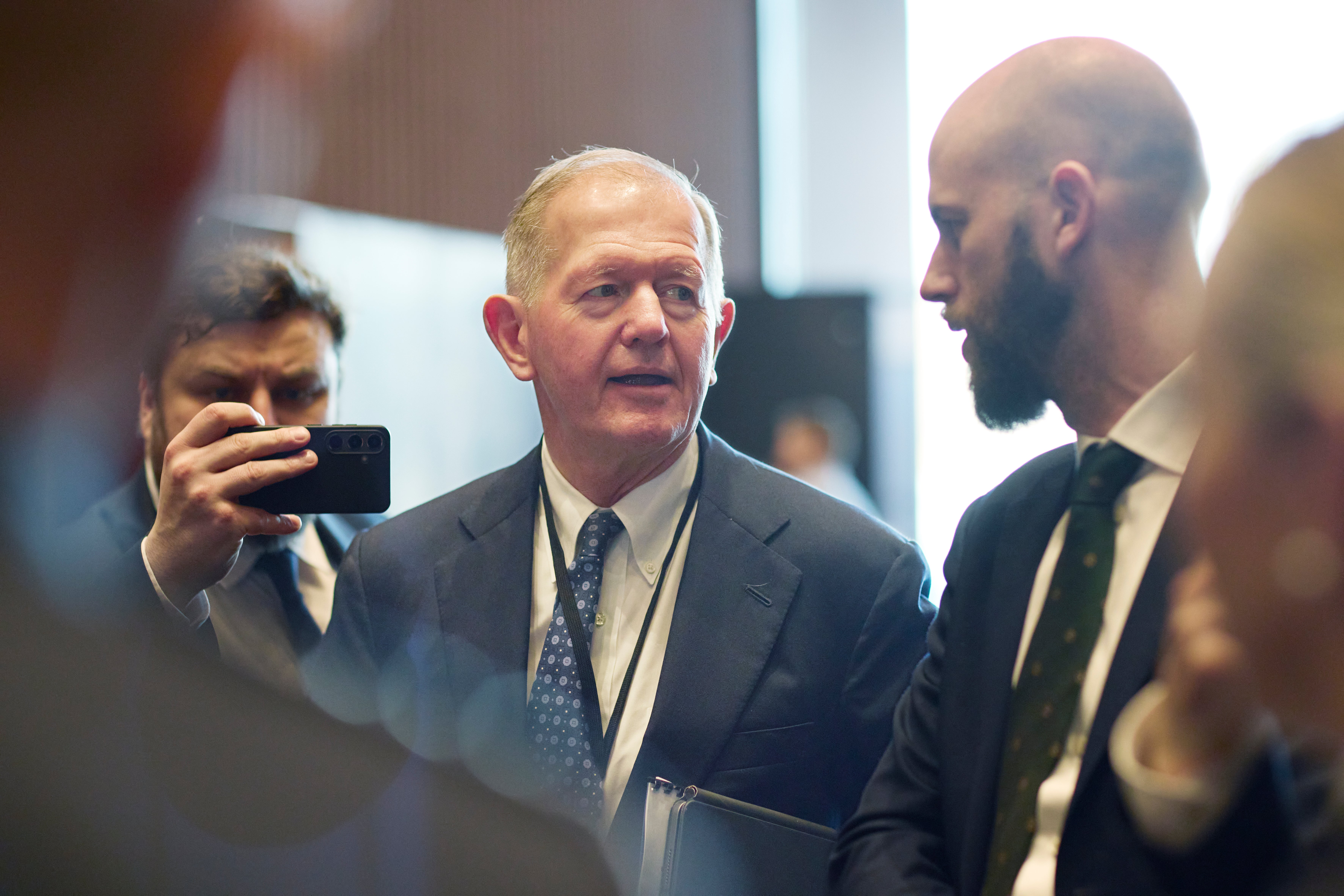 En man i mörkblå kostym och slips står bland andra män i formell affärsmiljö. En person håller upp en mobiltelefon.