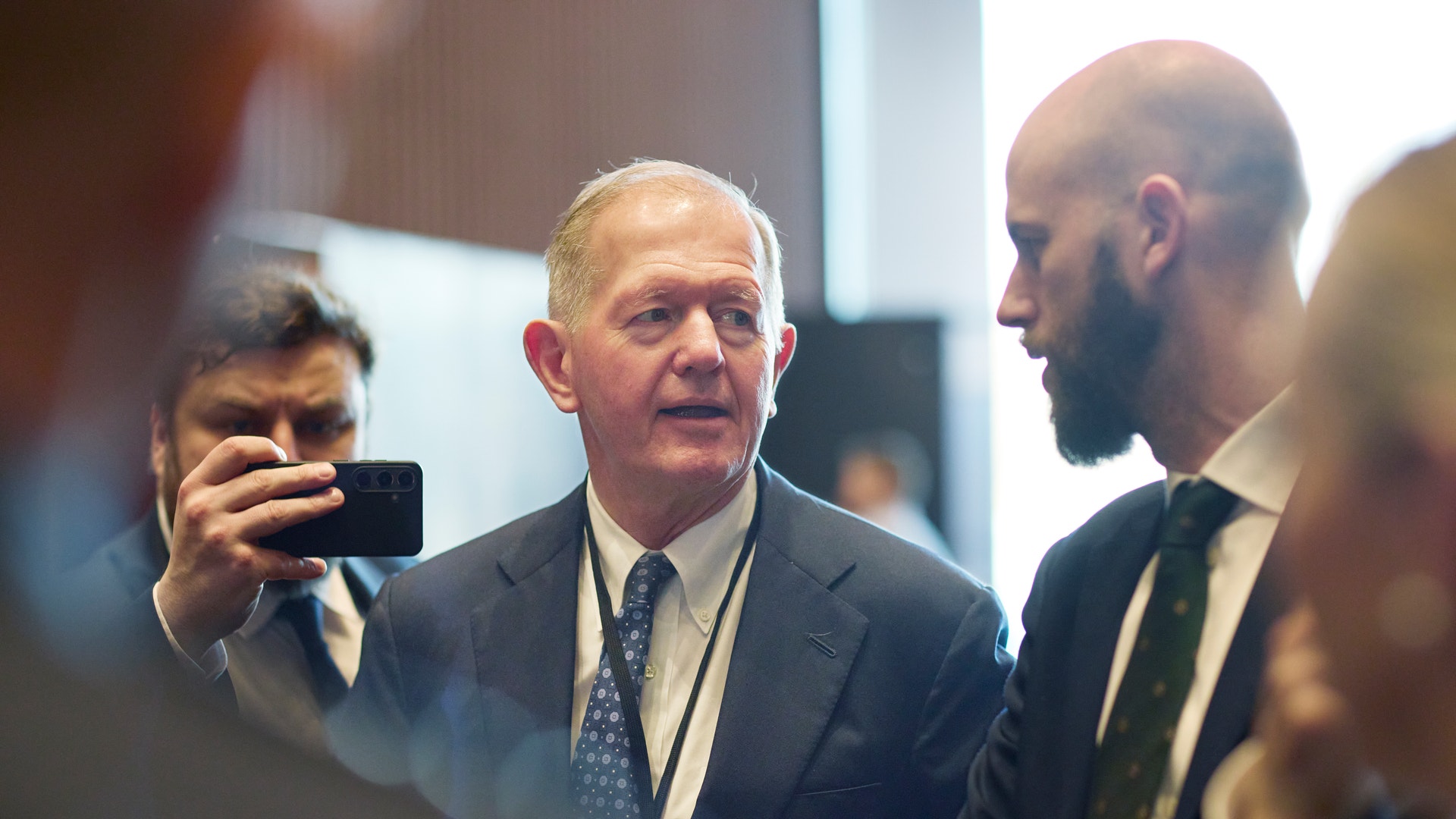 En man i mörkblå kostym och slips står bland andra män i formell affärsmiljö. En person håller upp en mobiltelefon.