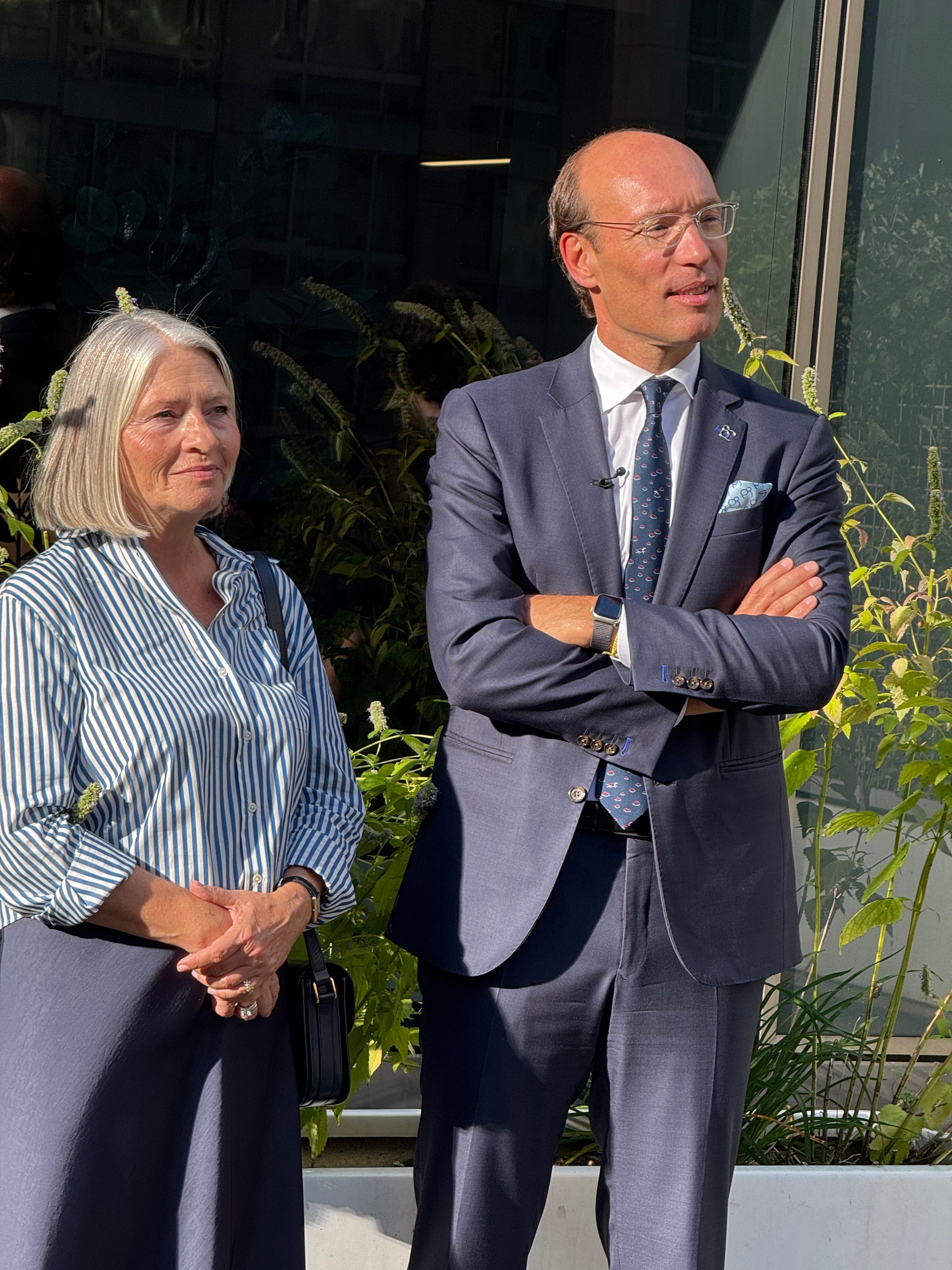 En kvinna och en man i kavaj står utomhus i solsken framför en byggnad med växter i bakgrunden.