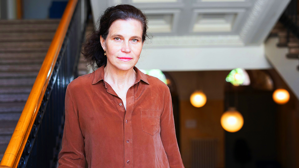 Porträttfoto av en kvinna vid namn Claudia. Hon bär en rostfärgad linneskjorta och står framför en modern arkitektonisk bakgrund med varma belysningsdetaljer. Hennes hår är uppsatt i en halv uppsättning, och hon tittar direkt mot kameran med ett lugnt uttryck.