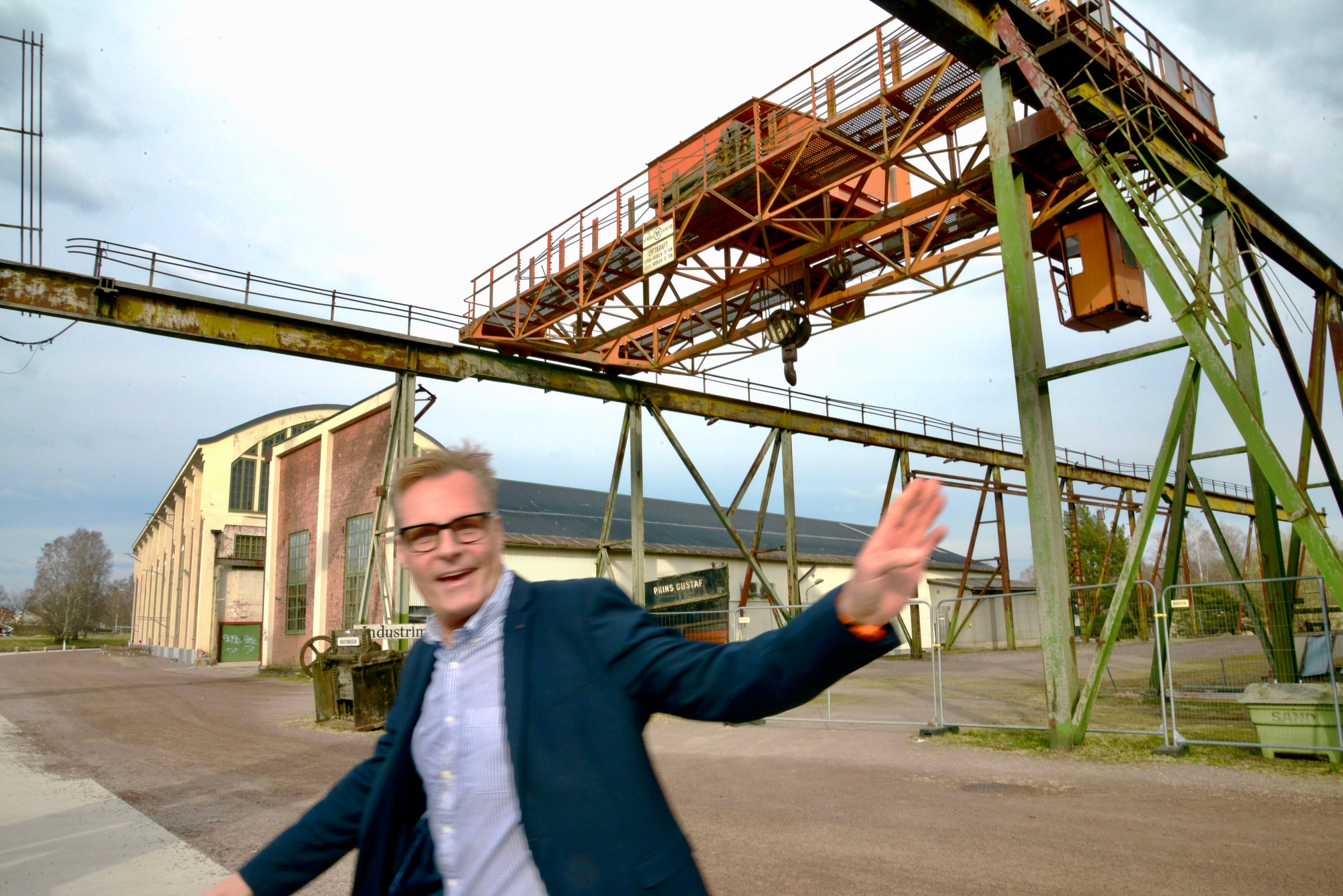 Man i kostym och glasögon gestikulerar framför en industrianläggning med kranstrukturer och tegelbyggnad.