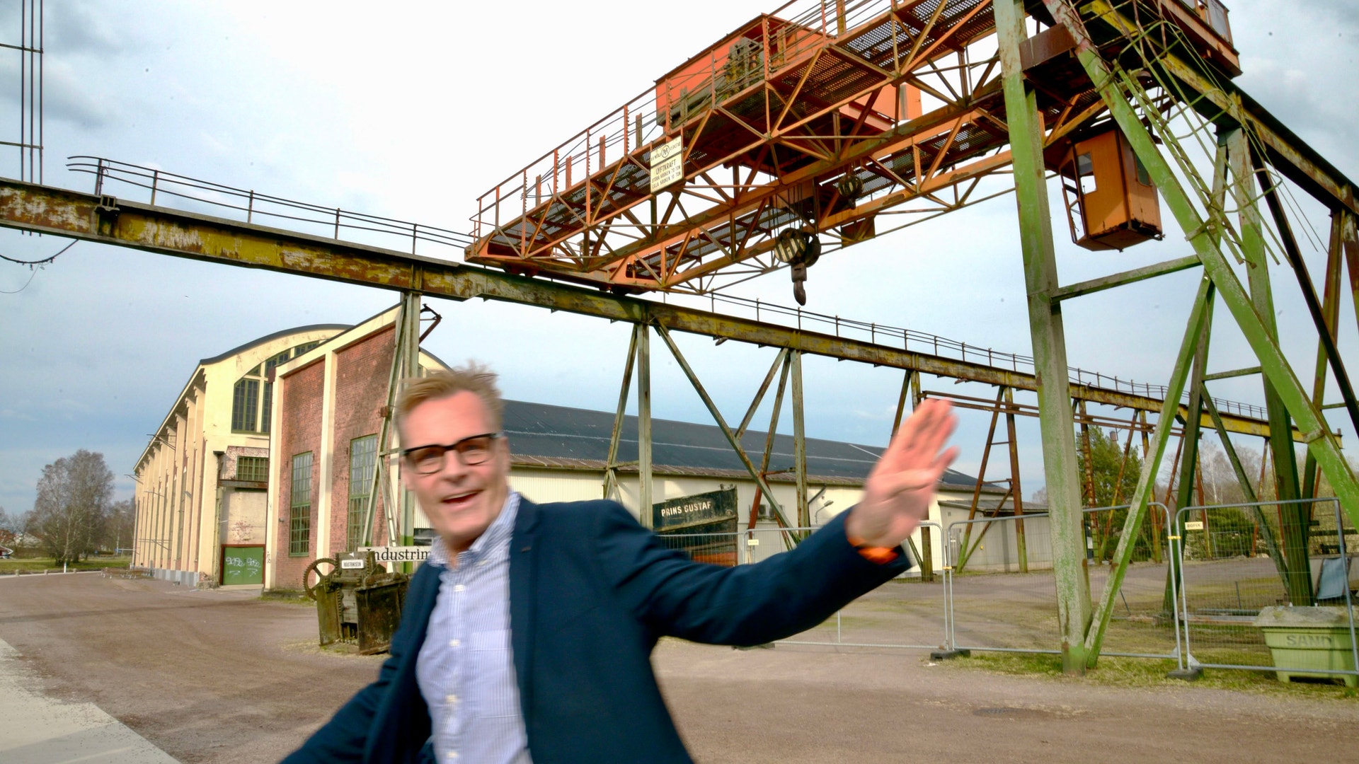 Man i kostym och glasögon gestikulerar framför en industrianläggning med kranstrukturer och tegelbyggnad.