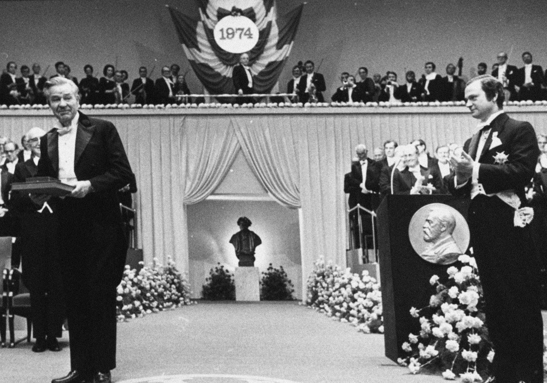 Svartvit fotografi från 1974 som visar en formell ceremoni med två män i smoking på en scen omgiven av blommor och åskådare.