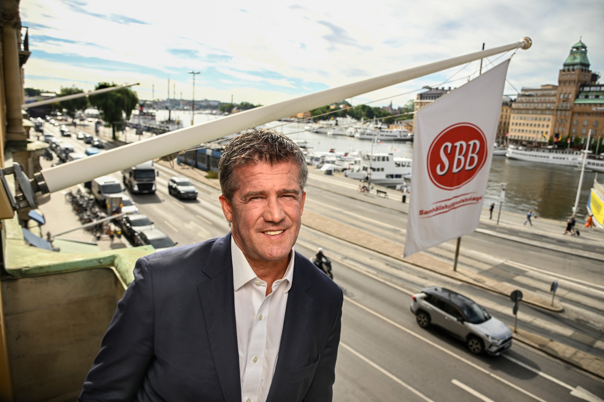 Leende man i mörk kavaj och vit skjorta står på en balkong med utsikt över en stadsgata och ett vattendrag. Bredvid honom hänger en flagga med SBB:s logotyp.