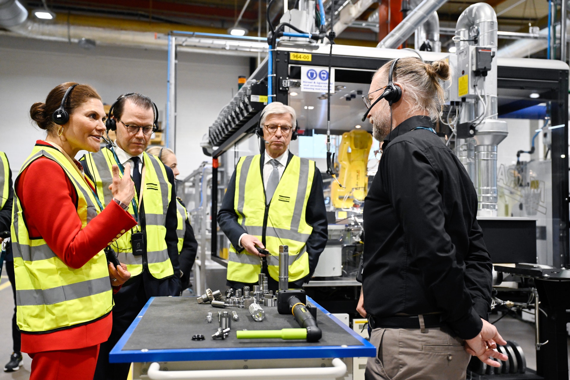 Flera personer i gula säkerhetsvärjar och hörlurar står i en fabrik och diskuterar omkring en industriell maskin på ett arbetsbord.