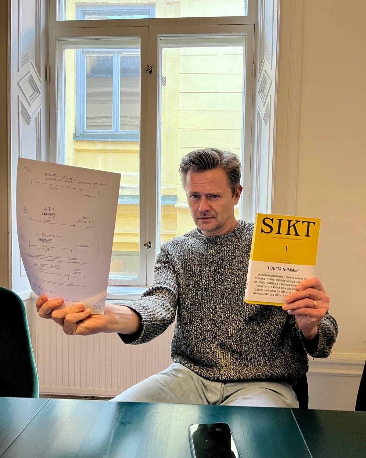 Man som håller upp en skiss och en gul bok med titeln SIKT vid ett bord