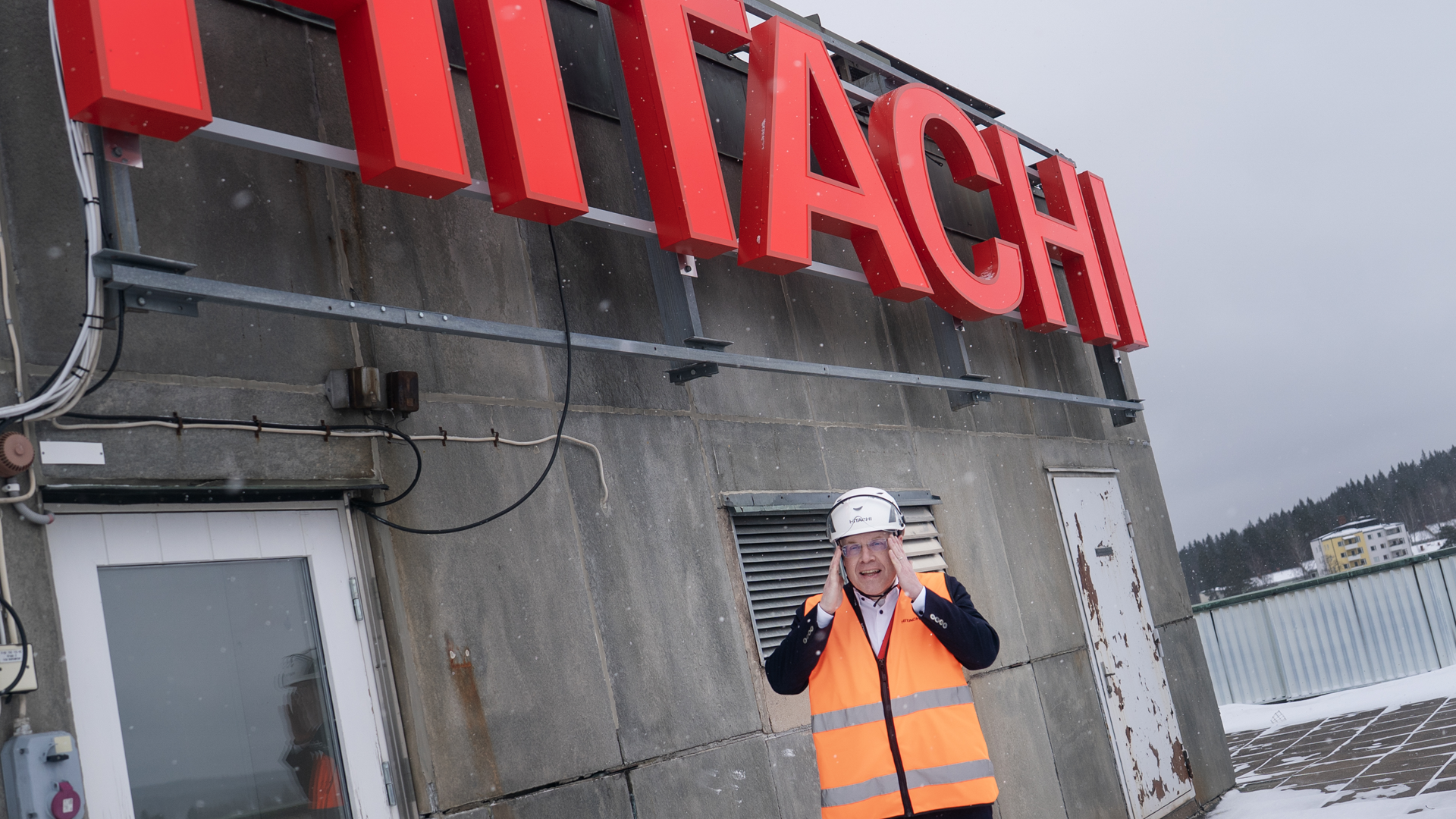 En man i orange säkerhetsvest och vit säkerhetshatt står framför en grå industribyggnad med röd Hitachi-skylt. Han ler mot kameran och är klädd i professionell arbetsutrustning. I bakgrunden syns snötäckt terräng och andra industribyggnader.