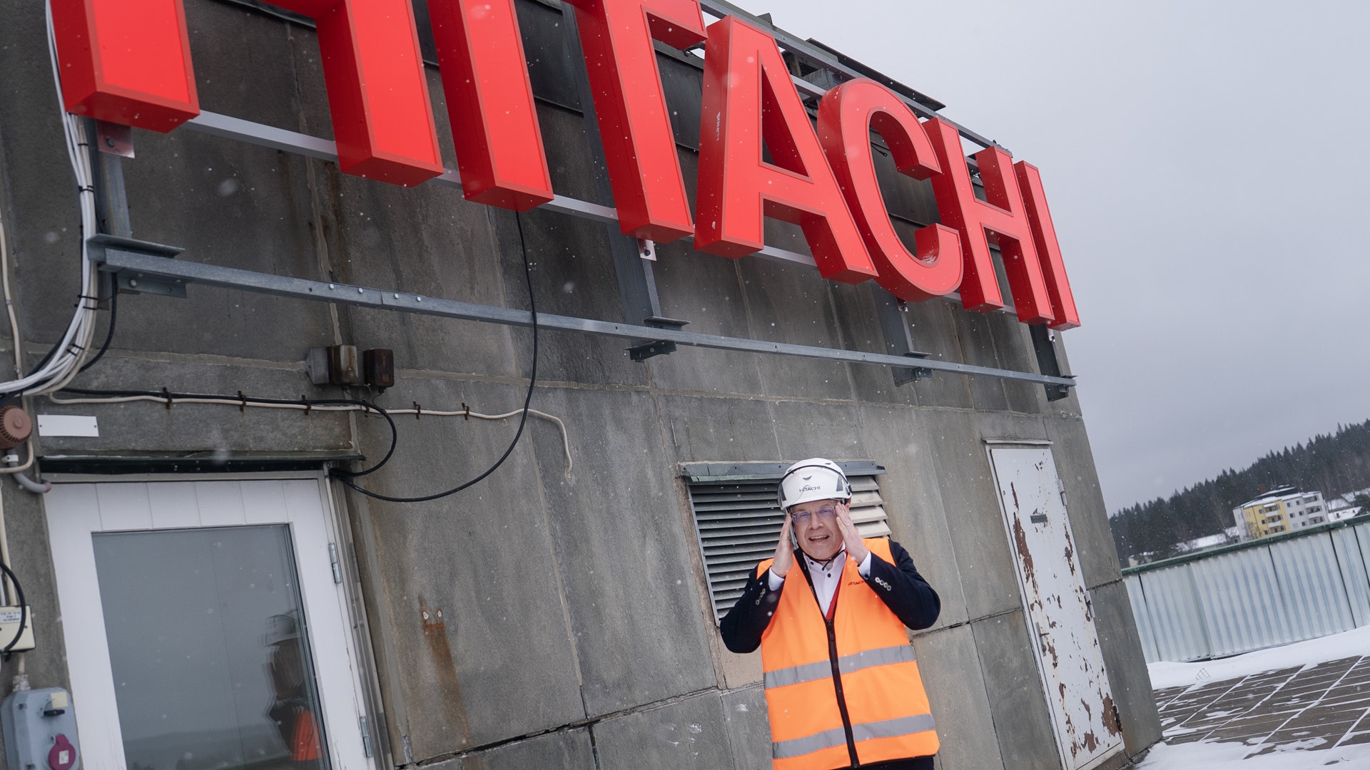 En man i orange säkerhetsvest och vit säkerhetshatt står framför en grå industribyggnad med röd Hitachi-skylt. Han ler mot kameran och är klädd i professionell arbetsutrustning. I bakgrunden syns snötäckt terräng och andra industribyggnader.