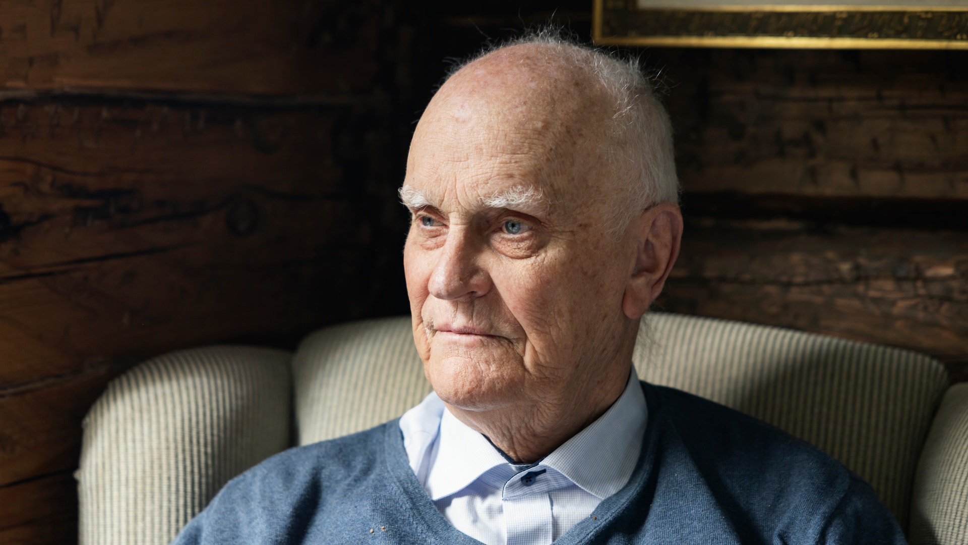 Porträtt av en äldre man som sitter i en stol. Han är klädd i en blå tröja och vit skjorta och tittar åt sidan.