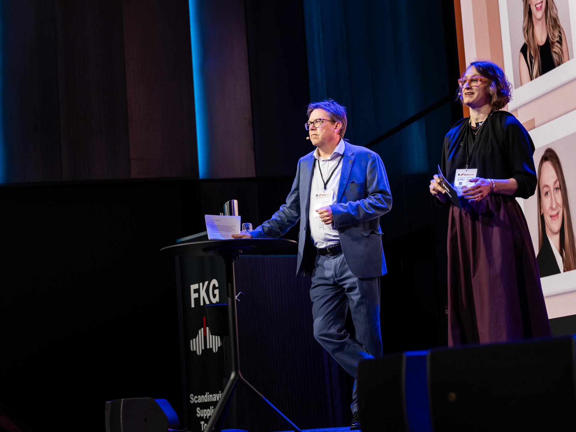 En man och en kvinna står på en scen vid en talarpodium med texten FKG och tittar mot publiken.
