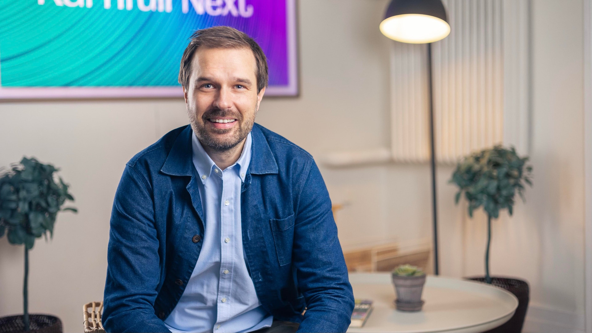 Porträtt av en man i blå skjorta och jeansjacka som sitter i ett modernt kontor.