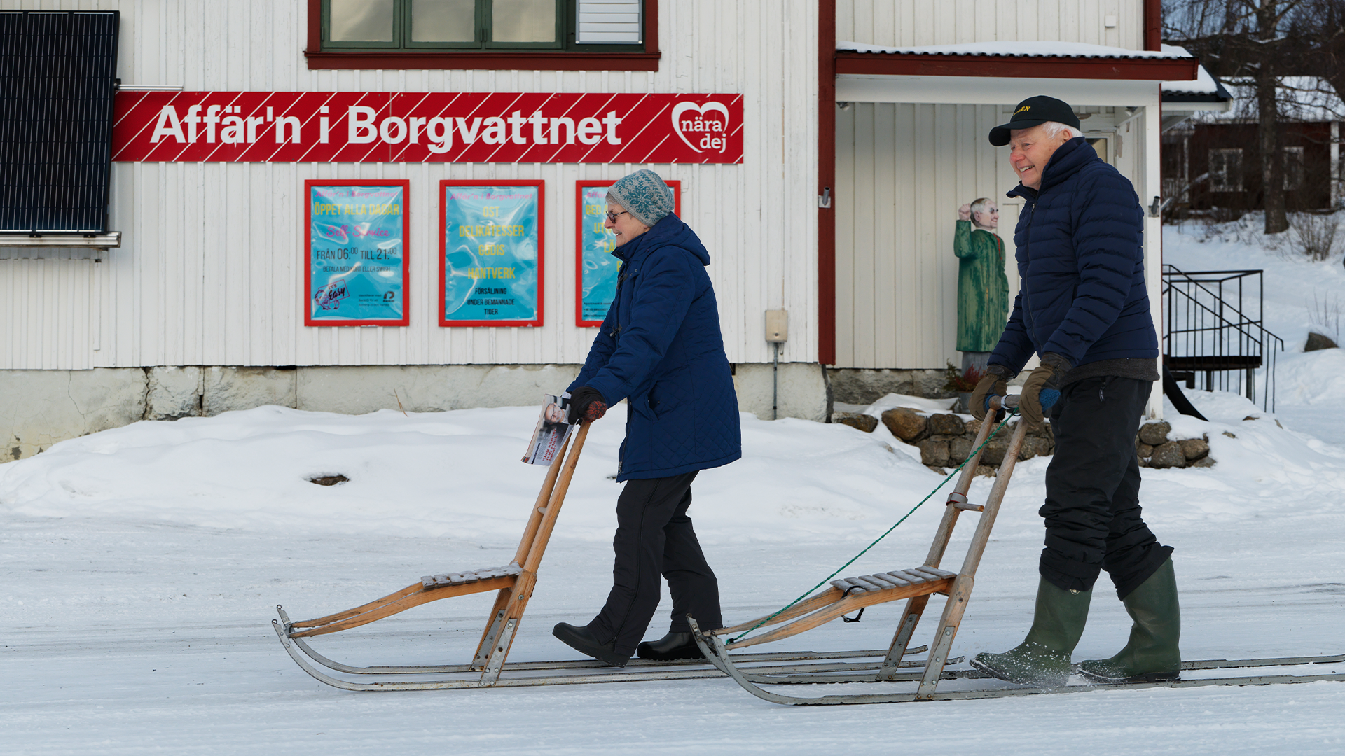 Två personer med träslädor i snön framför en röd byggnad