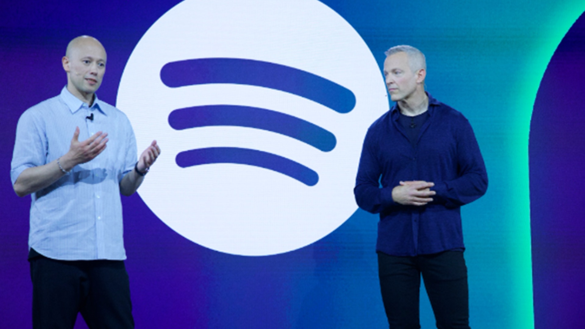 Två män presenterar framför Spotifys logotyp på en blå och turkos bakgrund.