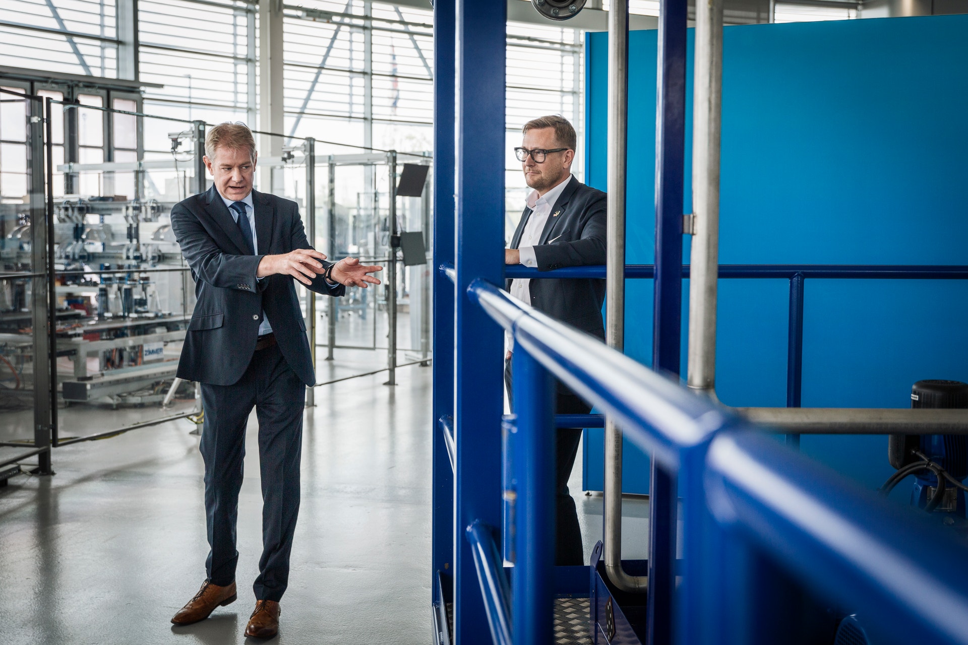 Två män i affärskläder diskuterar vid en blå vägg i en industriell fabriksmiljö.