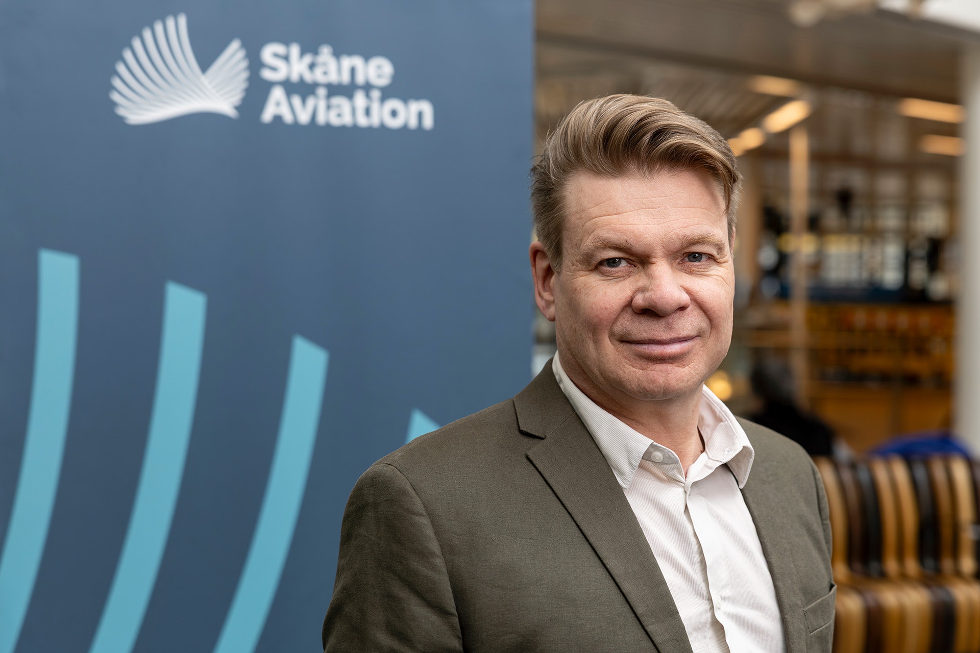 Man i grå kavaj och vit skjorta framför en blå Skåne Aviation-banner med vita vingar och ljusblå streck.
