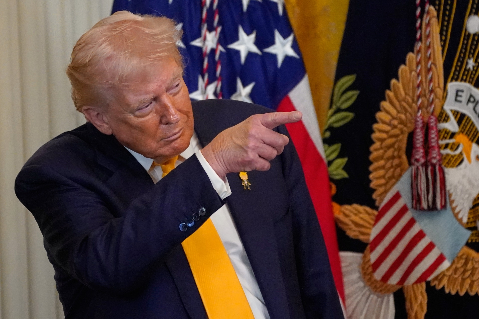 Man i mörk kostym pekar mot kameran framför amerikanska flaggor och officiella emblem.